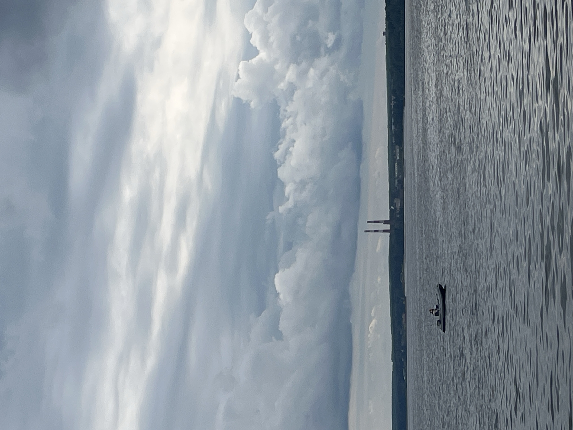 The image depicts a wide, grey body of water under a heavily clouded sky. In the foreground, the water is rippled. A small, dark inflatable boat with a white stripe is visible in the middle ground, containing a single person who appears to be fishing. In the distance, a dark green tree line runs along the horizon, beyond which two tall, red and white smokestacks stand out against the sky, indicative of an industrial area. The overall atmosphere is overcast and somewhat somber. This scene is likely the Kama River in or near Chaikovsky, Russia, given the presence of the smokestacks which are part of the Chaikovsky thermal power station.