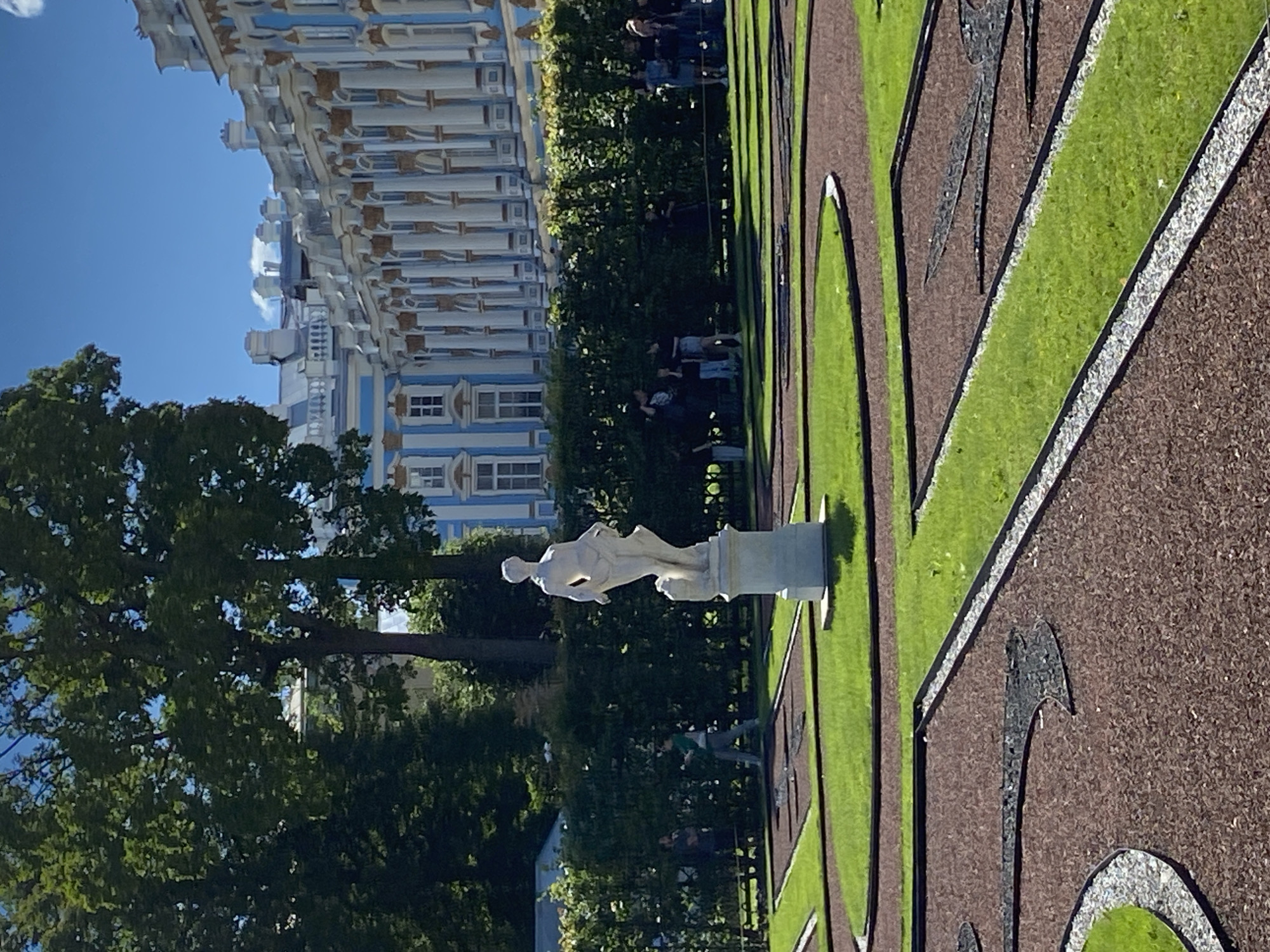 The image shows the Catherine Palace in Pushkin, Russia. In the foreground, a garden features manicured green lawns, pathways made of brown mulch, and decorative designs inlaid with dark material. A white statue of a woman stands on a pedestal in the center of a circular lawn. Behind the garden, a dense hedge forms a barrier, and beyond that, a crowd of people are visible, some standing and others walking. The palace itself, with its distinct blue and white facade and ornate columns, serves as the backdrop to the scene. The sky above is a clear blue with a few scattered white clouds. Trees with lush green foliage frame the left side of the image.