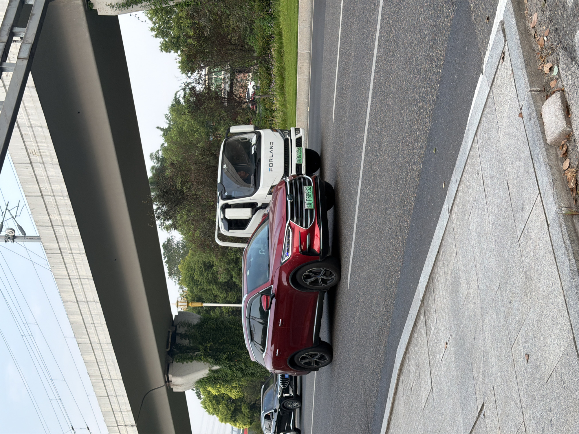 The image shows a red SUV driving on a road in Jiangjin District, Chongqing, China, under a raised highway structure. To the left of the red SUV, a black SUV is also visible. In the background, a white Forland truck is driving in the same direction. The road is asphalt with white lane markings. The raised highway structure is made of concrete and has electrical wires running above it. Lush green trees and vegetation line the right side of the road. The sky is overcast. The red SUV has license plate "A F68932". The white truck has license plate "A BZ9256". The scene appears to be a typical urban or suburban road with traffic.