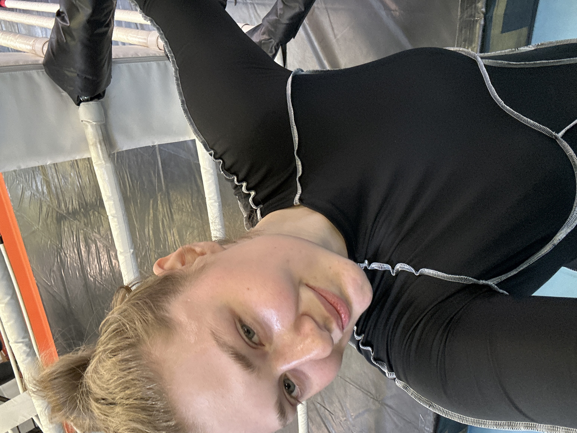 A close-up, tilted selfie of a light-skinned individual with blonde hair, partially styled in a bun. The person is wearing a close-fitting, long-sleeved black top with white edging along the seams. They have green eyes and light pink lips. The background appears to be a boxing or training ring, indicated by padded posts and barrier panels. The posts are white and gray with black padding. The backdrop includes gray panels and potentially orange elements. The location is unknown, though the prompt mentions Kazan, Russia.
