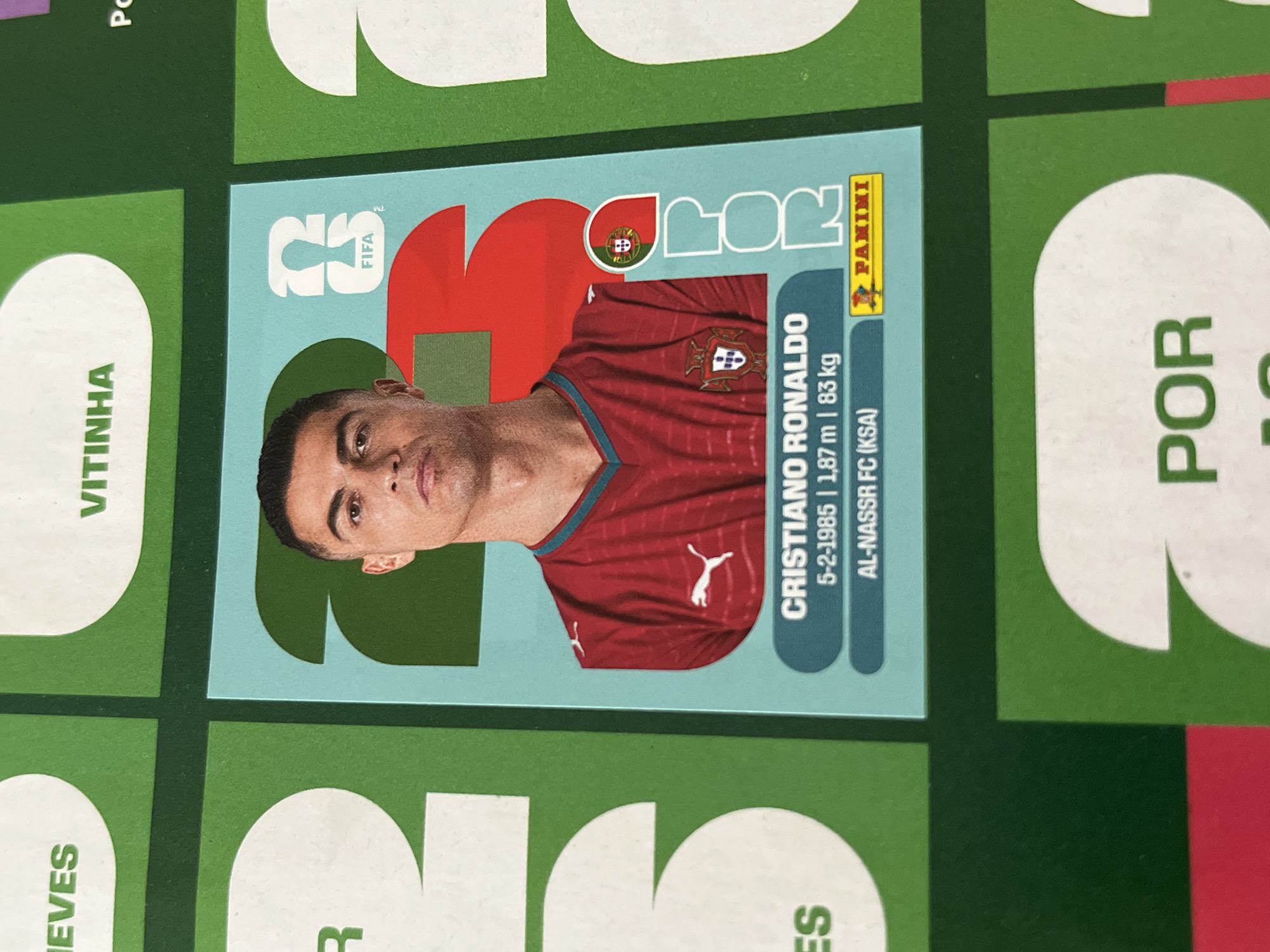 The image shows a FIFA Panini sticker featuring Cristiano Ronaldo. The sticker has a light blue background with the letters "POR" in a red and green color scheme above a portrait of Ronaldo. He is wearing a red Portugal jersey. Below the portrait, text reads "CRISTIANO RONALDO," followed by his birthdate (5-2-1985), height (1.87m), weight (83kg), and the name of his club (AL-NASSR FC (KSA)). The Panini logo is in the upper right corner. The sticker is surrounded by other partially visible stickers or album spaces. The other stickers feature the names "VITINHA", "NEVES", "POR", and other partially legible letters. The background behind the stickers is a dark green.