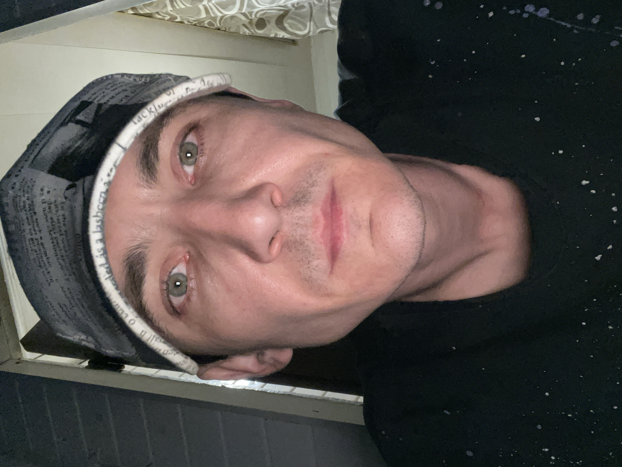A close-up selfie is taken of a person wearing a grey hat with a newspaper print pattern and a white band with text. The person has light skin and green eyes. They are wearing a black shirt with white speckles. The background appears to be indoors, with a patterned curtain visible at the top and what looks like a wall or door in the lower portion of the image. The lighting suggests it is either nighttime or an indoor setting with dim lighting. The person's head is tilted, and they are looking directly at the camera. The hat band has visible text, which reads "Laishevo is a lash...". The image has a slightly grainy quality.