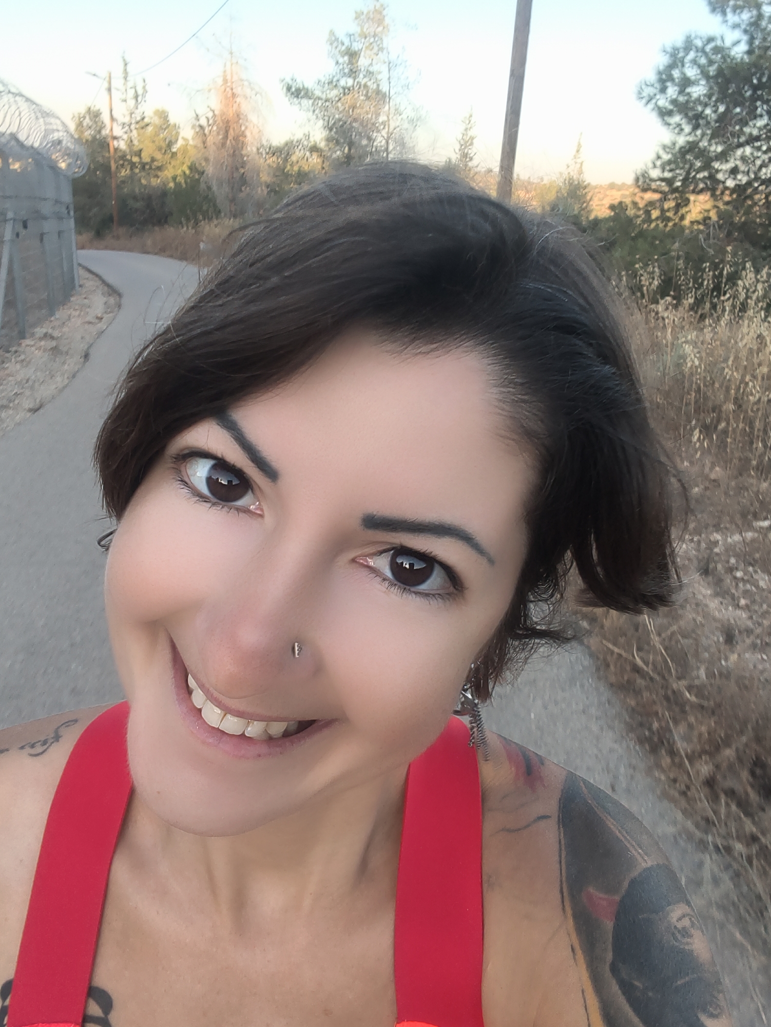 A medium shot photograph shows a woman with short, dark hair and brown eyes smiling at the camera. She is wearing a red top with thin straps, and a silver stud is visible in her nose. A large, dark tattoo covers her right arm and shoulder, and another smaller tattoo is visible on her left shoulder. She is outdoors, with a paved path and a wire fence visible behind her. The path curves to the left and appears to lead into a wooded area with dry grass and trees. The sky is a pale blue, suggesting either early morning or late afternoon light. The image appears to have been taken in Lachish Regional Council, Israel, based on the presence of a wire fence and arid vegetation characteristic of the region.