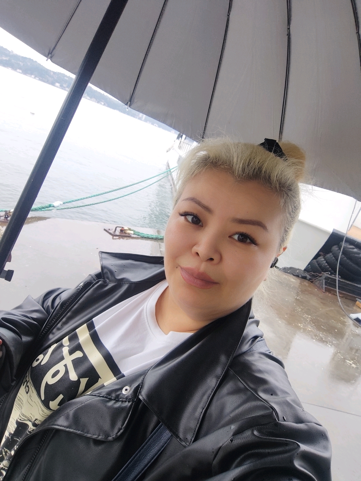 A selfie is taken from a slightly high angle of a woman standing under a gray umbrella outdoors. The woman has blonde hair pulled up in a bun with a black ribbon, and is wearing a black leather jacket over a white t-shirt with black and yellow graphics. She has on makeup and is looking at the camera. In the background, there is a body of water, a pier with ropes and cleats, and a distant hillside with buildings, suggesting the location is in Sarıyer, Turkey, likely near the Bosphorus strait, given the presence of water and boats. The ground appears wet, indicating it may have rained or is raining.