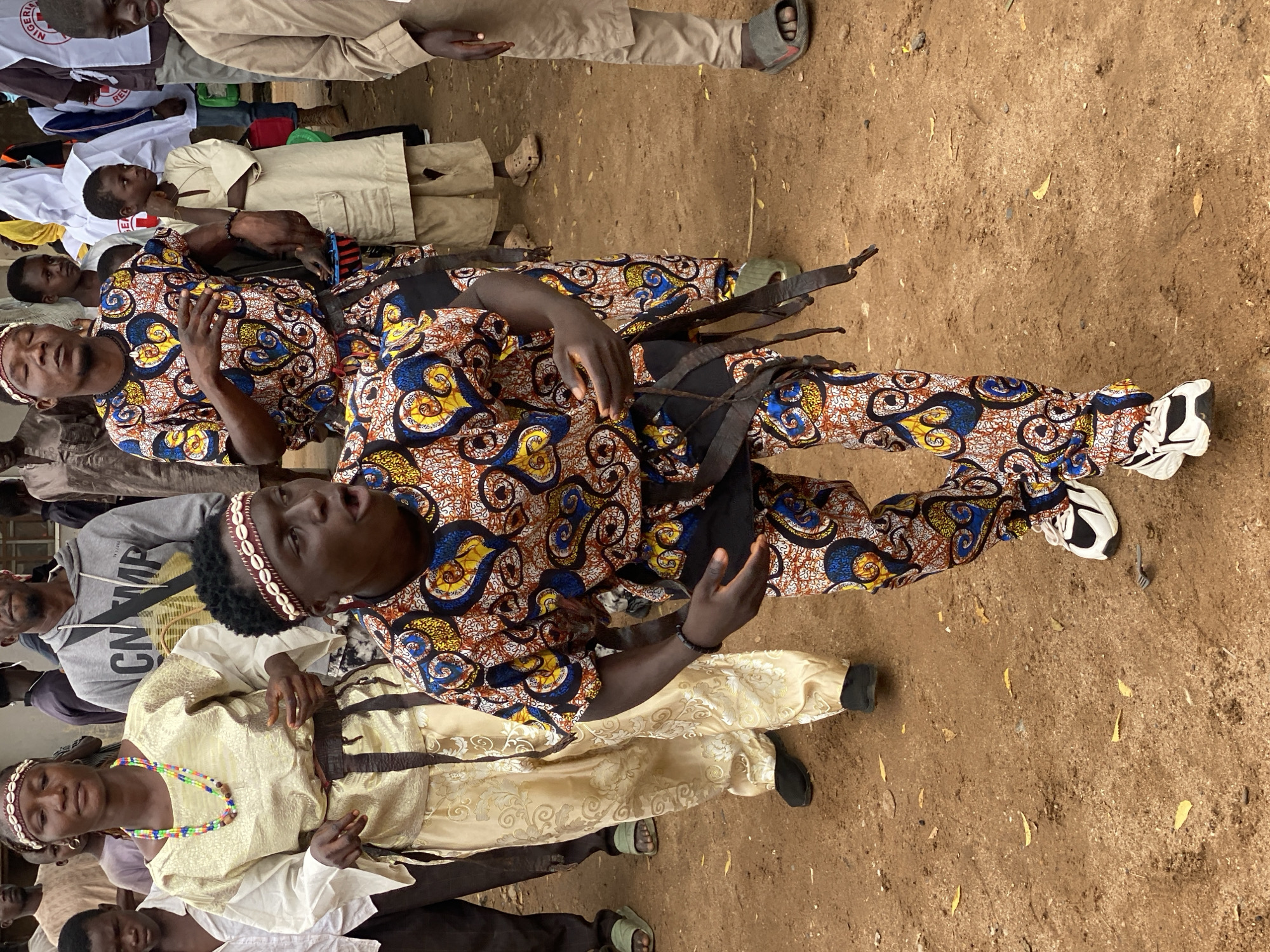 A group of people, some in traditional attire, stand on a dirt ground in Rigachikun, Nigeria. A young man in the foreground, wearing a colorful patterned outfit and a beaded headband, appears to be dancing or performing, with his mouth open and hands outstretched. Behind him, a woman in a golden traditional dress and beaded necklace stands with her eyes closed. Other individuals, including children and adults, are visible in the background, some wearing more modern clothing like a grey hoodie and others in white or beige garments. The environment is outdoors, with dry, dusty ground and scattered leaves.