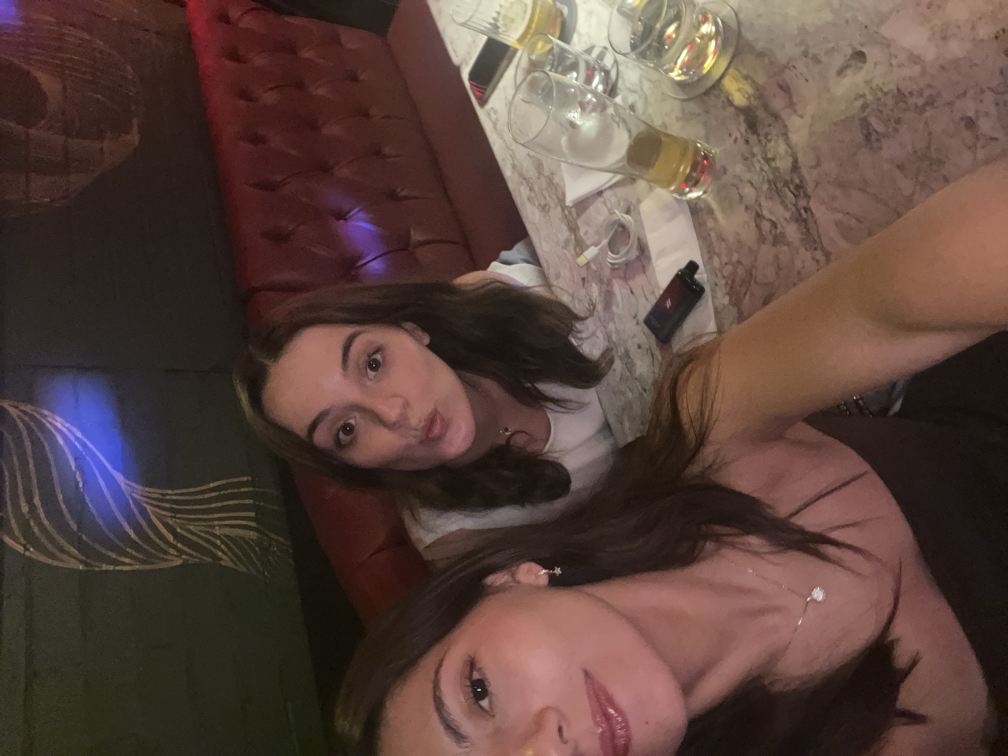 Two women are posing for a selfie at a bar or restaurant. There is a red leather booth seat, and a marble-looking table with drinks and a charging cable. A dark green patterned wall is visible to the left. The location is likely in Puebla, Mexico.