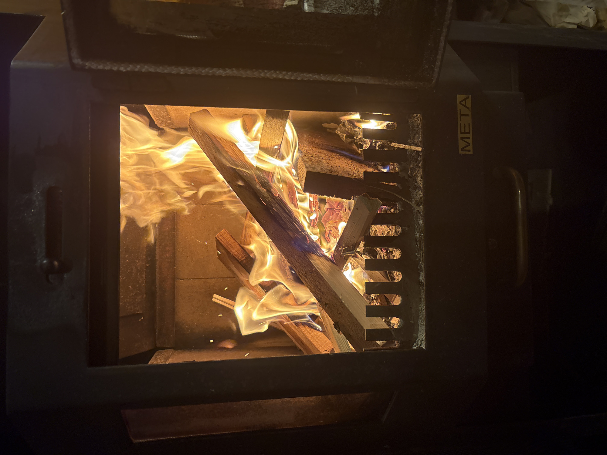 The image shows a close-up view of a fireplace with burning logs and flames. The fireplace appears to be made of dark metal with a glass door partially open. Inside, there is a grate at the bottom, and the walls of the fireplace are made of brick or stone. A yellow label with the word "META" is visible on the front of the fireplace. The background is dark, suggesting the photograph was taken in a dimly lit room. There are no people visible in the image, and the scene depicts a typical domestic fireplace in use. The location in Lyady, Russia is not discernible from the visual information present in the image.