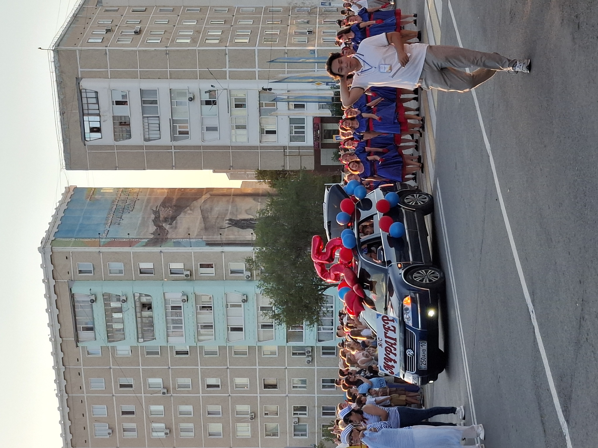 A group of people are gathered on a street, possibly for a parade or celebration. In the foreground, a dark-colored Volkswagen Passat is adorned with a large "25" made of red balloons, along with other red and blue balloons. A banner on the car reads "Я/С МУРАГЕР", indicating a name or organization. Several people, including women in blue dresses with red trim and men in casual attire, are visible near the car and on the sidewalk. Behind them, two tall, multi-story apartment buildings with many windows dominate the background. The sky is a pale orange, suggesting it is either sunrise or sunset. The street is paved and has white lane markings. A young man in a white polo shirt and grey pants walks towards the camera on the right side of the frame.