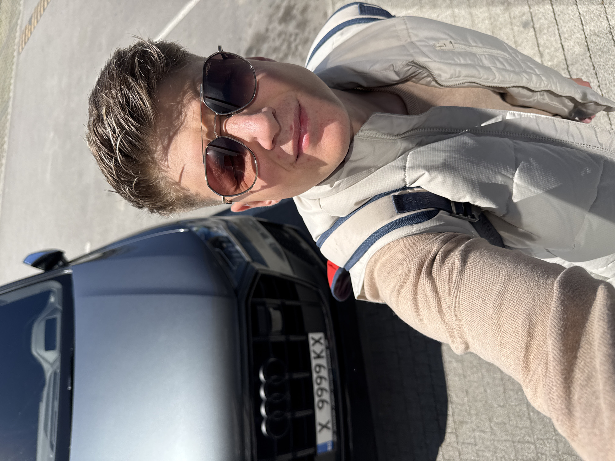 A person stands next to a gray Audi car, likely in an outdoor parking area. The person is wearing sunglasses, a beige sweater, and a light-colored vest. The license plate of the Audi reads "X 9999 KX". The background appears to be a paved surface under bright sunlight. It is not possible to definitively identify the exact location within Plovdiv, Bulgaria without further information, but the presence of the car with a Bulgarian license plate suggests the location is within Bulgaria.