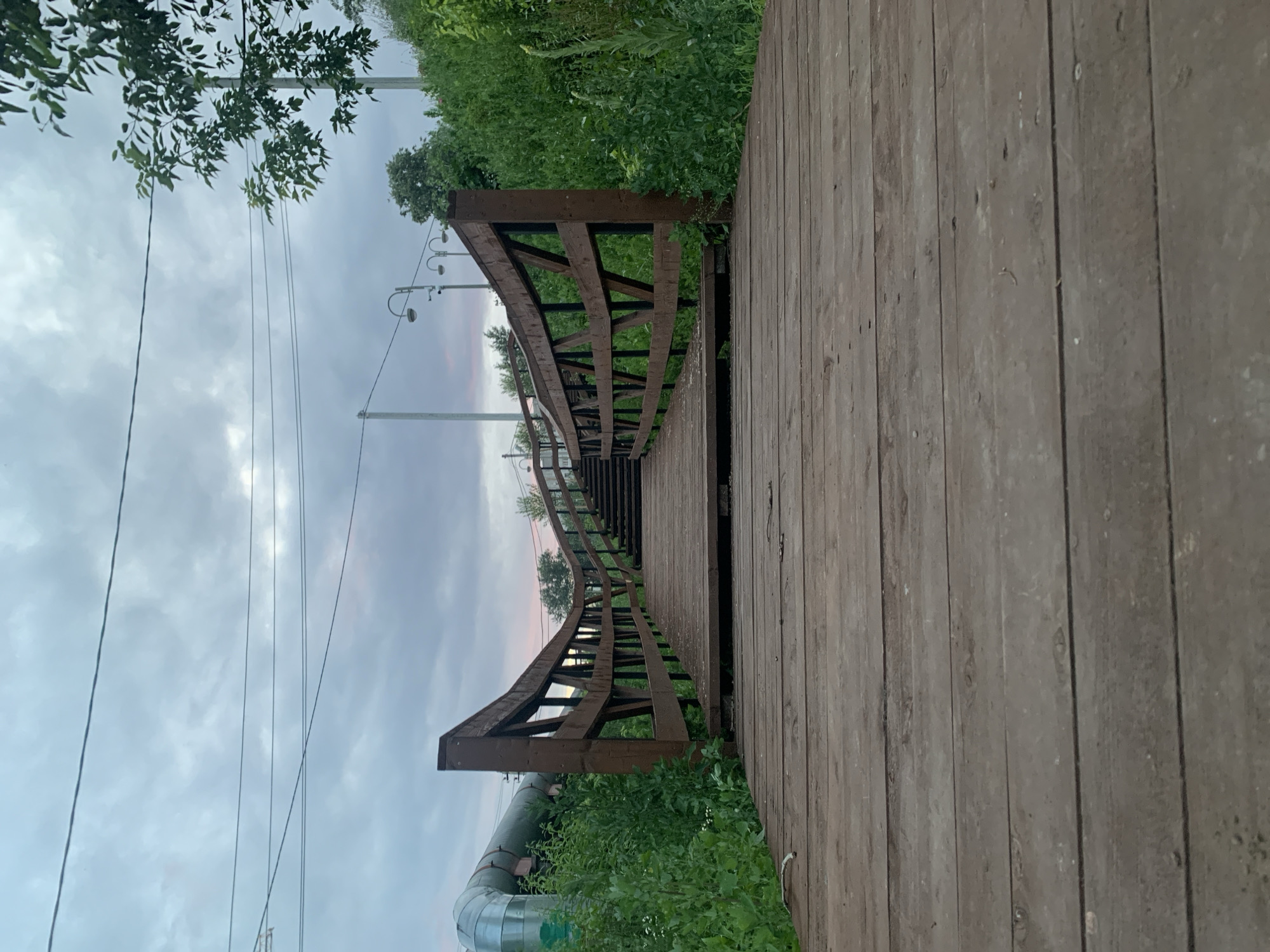 The image shows a wooden bridge with railings leading up a set of stairs. The bridge is made of weathered wooden planks and has a dark brown wooden railing with a crisscross pattern. Lush green vegetation surrounds the bridge and stairs, with a large metal pipe visible on the left side. In the background, there are streetlights and power lines against a cloudy sky with hints of pink from a sunset or sunrise. The perspective is low, looking up the bridge from ground level. This scene is located near the Kama River embankment in the city of Perm, Russia.