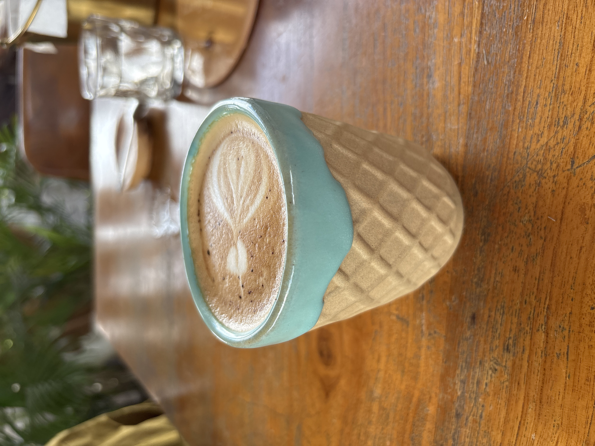 A close-up, top-down shot captures a cup of coffee with latte art placed on a wooden table. The ceramic cup is light beige with a textured, waffle-like pattern on its body. A portion of the cup's rim and upper section is glazed in a smooth, light turquoise or teal color, with an organic, flowing edge where it meets the beige body.
Inside the cup, the coffee, likely a latte or cappuccino, features distinct white foam with a heart-shaped latte art design. Small, dark speckles, possibly coffee grounds or cocoa powder, are visible on the foamed milk.
The wooden table surface, which serves as the primary background, exhibits visible grain patterns and areas of wear or discoloration, suggesting age or frequent use. In the blurred background, to the upper left of the cup, a translucent glass object is partially visible, along with hints of green foliage from a plant on the far left. The lighting appears natural, suggesting it is daytime. The overall scene evokes a calm and relaxed atmosphere, likely in a cafe or similar setting in Kuta Selatan, Indonesia. No discernible text is visible in the image.