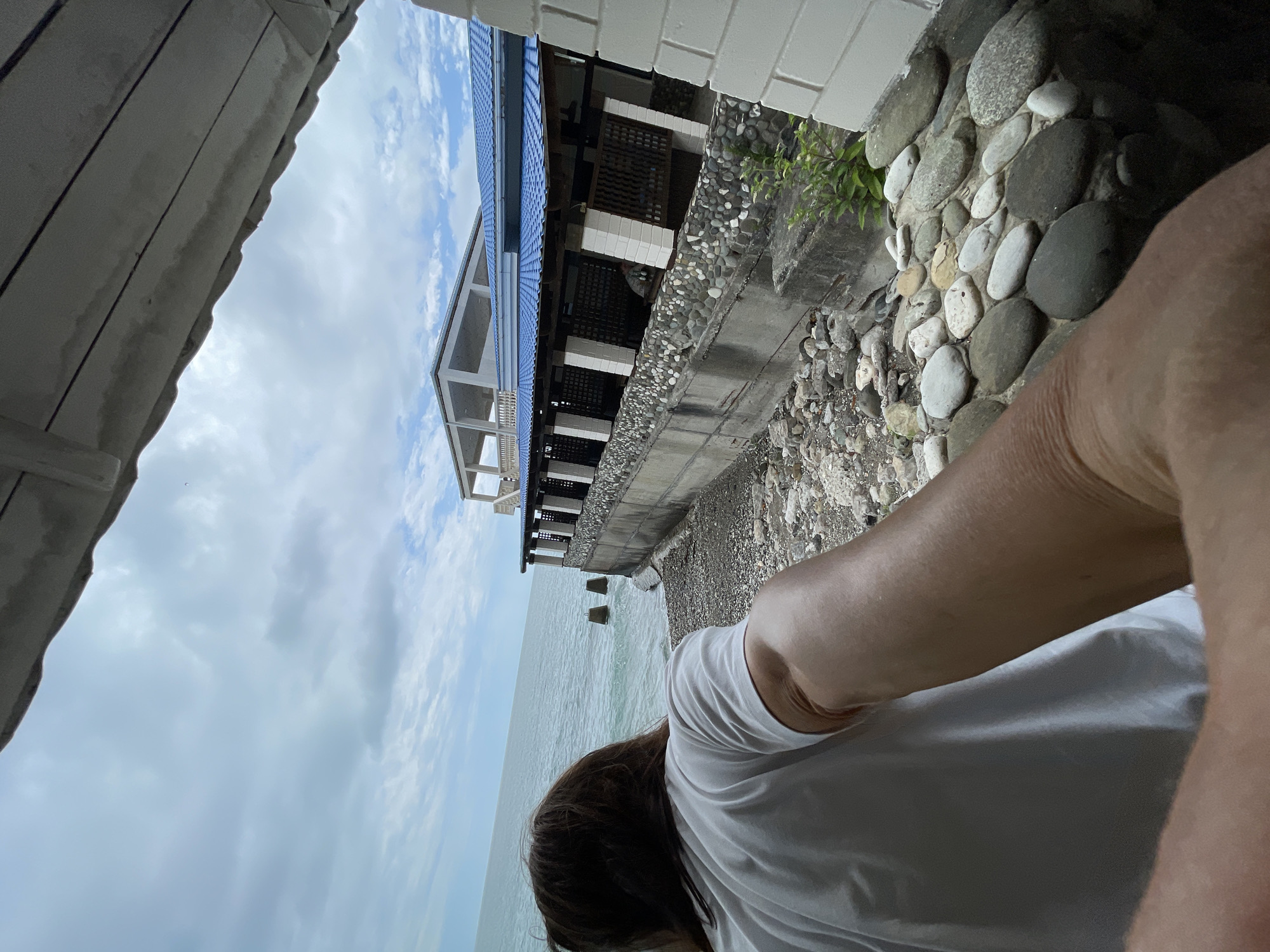 An over-the-shoulder, wide-angle shot captures a person in a white t-shirt looking out at the sea from a covered porch in Abaata, Abkhazia. The person's right arm and hand are extended towards the camera, obscuring the lower right corner. The sea, a tranquil blue-green, stretches to the horizon under a cloudy sky. Two weathered wooden posts stand in the water near the shore. A building with a blue tiled roof and open-air covered walkways is situated on a raised concrete embankment overlooking the sea. The embankment is lined with a wall constructed of grey concrete and large, smooth, multi-colored river stones. The ground leading down to the sea consists of similar stones and some gravel. The scene is set during the day, with overcast conditions.