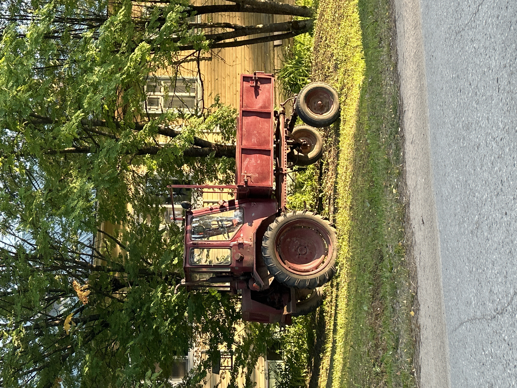 A weathered, dark red tractor with a trailer attached is parked on a grassy shoulder next to an asphalt road. The tractor is positioned diagonally, with its large rear wheel prominent in the foreground. The trailer is box-shaped and also dark red. Behind the tractor, a light-colored wooden building with windows is visible, partially obscured by lush green trees. The scene is bathed in sunlight, suggesting a bright, clear day. The grass on the shoulder is green and appears well-maintained. The overall impression is one of a rural or semi-rural setting, possibly a farm or a country road. There are no people or animals visible in the image. The image is taken from a slightly elevated angle, looking down at the tractor and the road. The time of day appears to be late afternoon, based on the angle of the shadows cast by the trees.