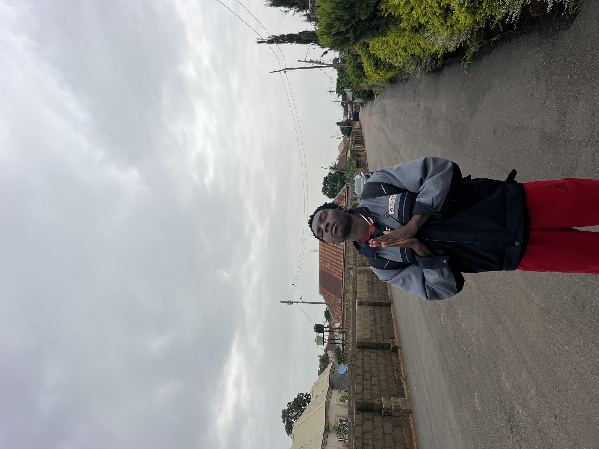 A young Black person stands in the middle of a paved road, with their eyes closed and hands clasped together in front of their chest, as if praying or meditating. They are wearing a blue and gray jacket over a red garment, and red pants. A backpack is visible on their back. The road is lined with brick walls and hedges, with houses and trees visible in the background. The sky is overcast and cloudy.