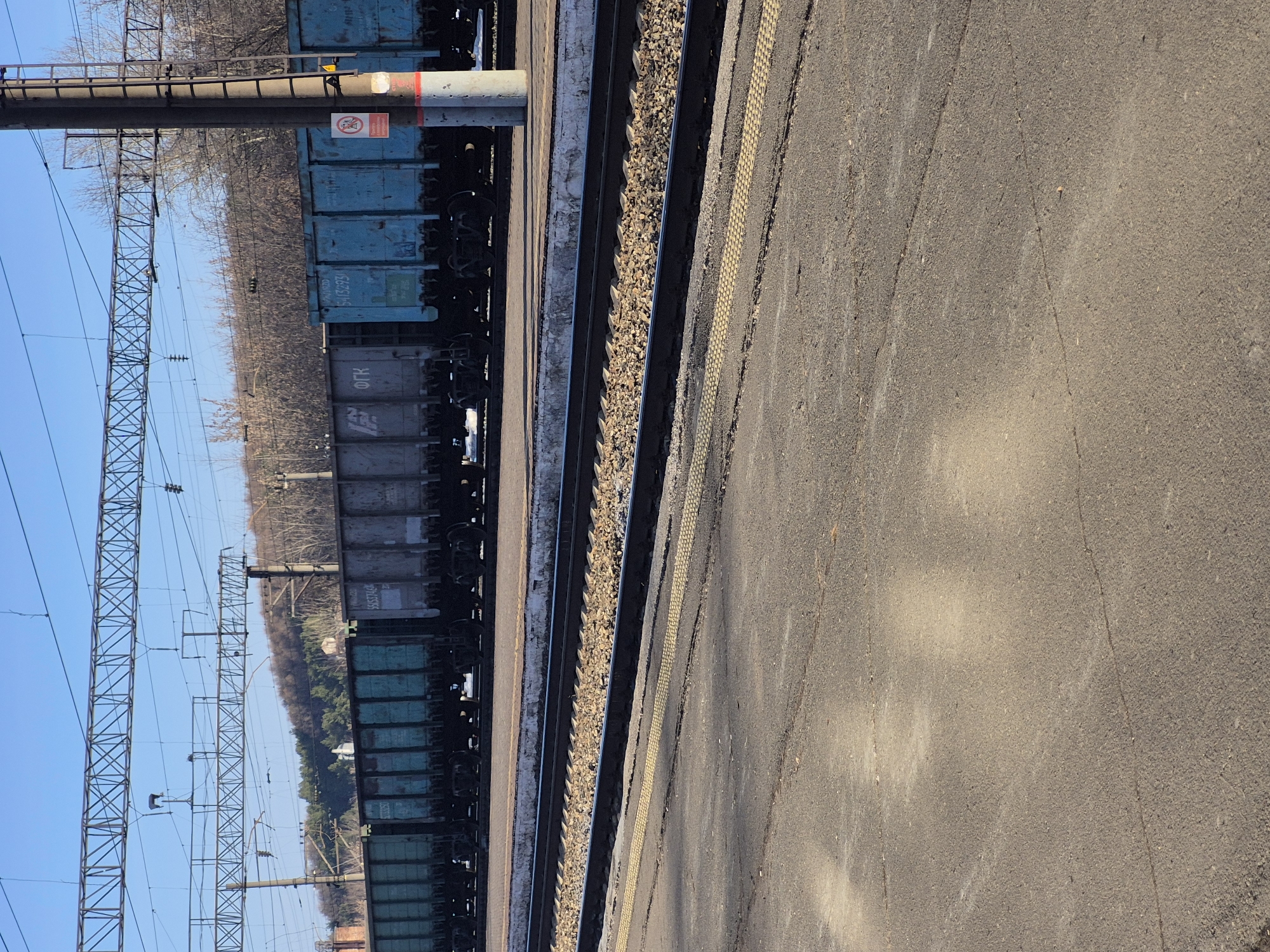 The image depicts a railway scene, likely in the city of Neftekamsk, Russia. A series of freight train cars, predominantly blue and gray, are stationary on the tracks. Parallel to the train tracks is a concrete platform marked with a yellow safety line. Overhead, a network of power lines and supporting pylons are visible against a blue sky. In the background, beyond the railway, there's a line of trees with bare branches. The general environment suggests an industrial or transportation area. No people are visible.