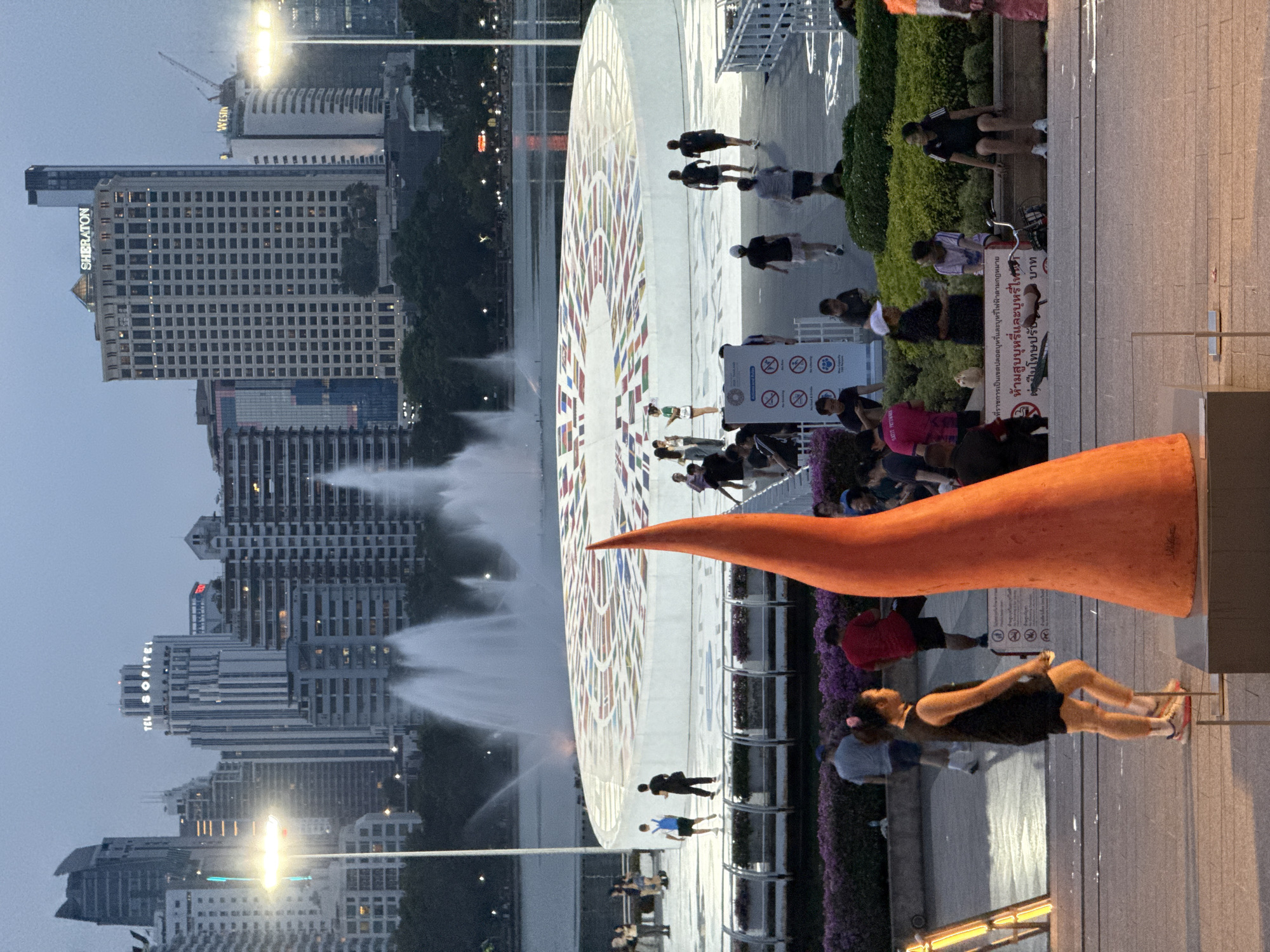 The image depicts a scene in Bangkok, Thailand, featuring the Queen Sirikit National Convention Center and surrounding cityscape at dusk. In the foreground, a bright orange, conical sculpture stands prominently on a paved area. Various people are walking around the plaza, some near the sculpture and others around a large, circular structure, possibly part of an event. To the left, a water fountain display is visible. The cityscape in the background includes several tall buildings, with recognizable brands such as Sheraton, Westin, and Sofitel visible on the buildings. Signage with Thai script is also present.