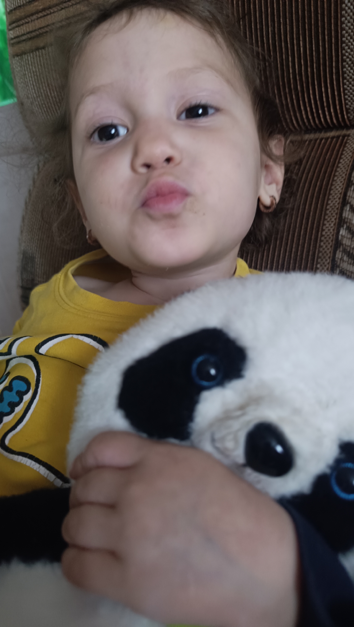 A close-up, over-the-shoulder shot features a young child holding a stuffed panda toy. The child, positioned in the foreground, looks directly at the camera with puckered lips and dark eyes. Their brown hair is slightly messy, and they are wearing a yellow t-shirt with a graphic design. A small, brown hoop earring is visible on their right ear. The stuffed panda is held close to the child's face, obscuring part of their body. The background consists of a textured, brown sofa. The scene appears to be indoors, likely in a residential setting. There is no indication of the child's specific location in the city of Karsha, Russia.