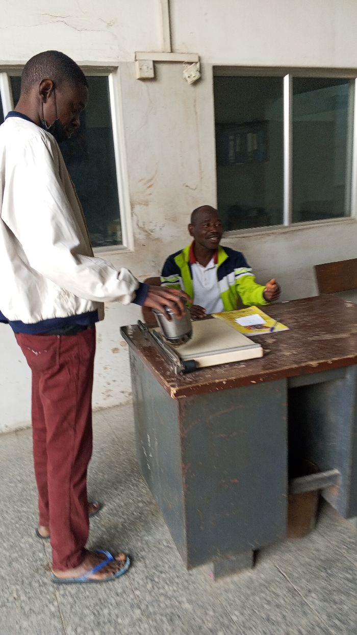 The image captures an indoor scene in Dakata, Nigeria, featuring two adult males interacting around a desk. A male subject stands on the left, clad in a light-colored jacket, maroon trousers, and blue-patterned flip-flops. He wears a black face mask lowered beneath his chin and is actively pressing a silver, cylindrical object onto a grey paper cutter or laminator situated on the desk. His attention is focused downward on this task. A second male subject is seated behind the desk to the right, wearing a white polo shirt layered with a green, blue, and white jacket. He is smiling and looking towards the standing individual, with his right hand resting on the dark brown, heavily worn wooden desk surface, near a yellow notebook and a pen. The desk itself has a dark grey base. The background consists of a light-colored wall exhibiting stains and cracks, with multiple rectangular windows featuring dark frames. A white electrical light switch and a damaged fixture are visible on the wall above the windows. The floor is tiled in grey. The overall environment suggests an operational, possibly public or administrative, space showing signs of age and use.