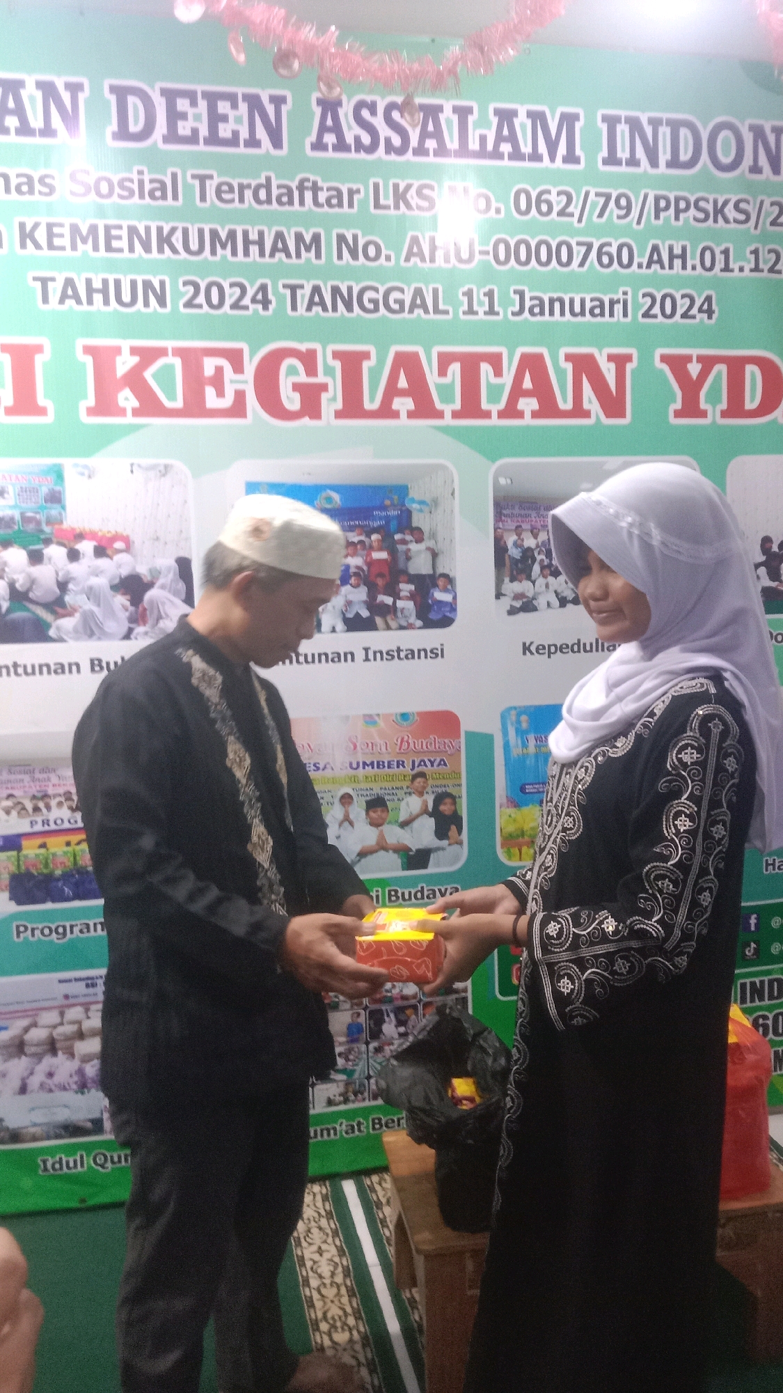 The image captures an indoor event, likely a community gathering or celebration, in Sumberjaya, Indonesia. The background features a large banner with text, partially obscured by individuals. The banner appears to be an official announcement or advertisement, with prominent Indonesian script and numbers. Dominating the scene are two individuals, a man and a woman, in the foreground. The man, wearing a black traditional outfit and a white skullcap, is seen handing a small, yellow box to the woman. The woman, dressed in a black hijab and a matching outfit adorned with intricate white patterns, is accepting the box. Their interaction suggests a gift exchange or presentation. To the right of the woman, a black plastic bag lies on the floor, potentially containing other items. The overall lighting is moderate, highlighting the subjects and the banner. The floor has a patterned carpet.