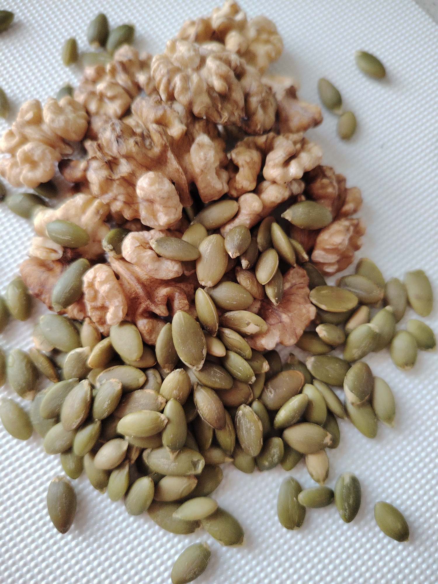This is a close-up, top-down shot of a pile of walnuts and pumpkin seeds on a white textured surface. The walnuts are a light brown color and have a wrinkled, irregular shape, appearing to be shelled. The pumpkin seeds are a muted green color and are elongated and flat. They are scattered amongst and around the walnuts, with some appearing to be partially covered by the walnuts. The lighting is bright and even, suggesting it might be daytime with diffused light. The background is a white, woven-like material that provides a neutral backdrop, allowing the subjects to stand out. No people, text, or specific location cues are visible in the image. The overall impression is a simple arrangement of healthy food items.