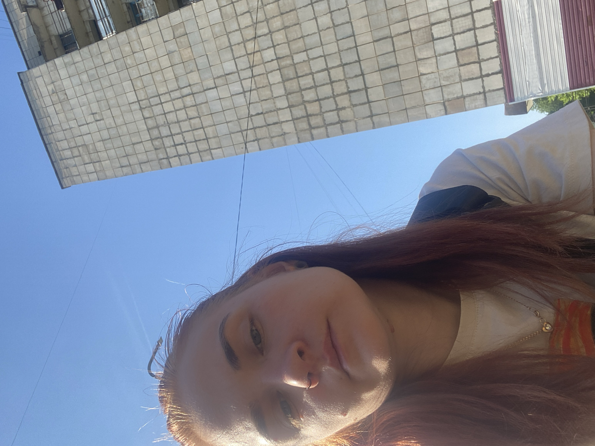A low-angle selfie shows a red-haired woman against a blue sky. Behind her, a large building is visible, its facade comprised of numerous square tiles. Based on the building’s architecture, this photo was taken in the city of Chaikovsky, Russia. The woman is wearing a white shirt with a dark jacket on her shoulders and a delicate necklace. The sunlight is strong, causing some lens flare.