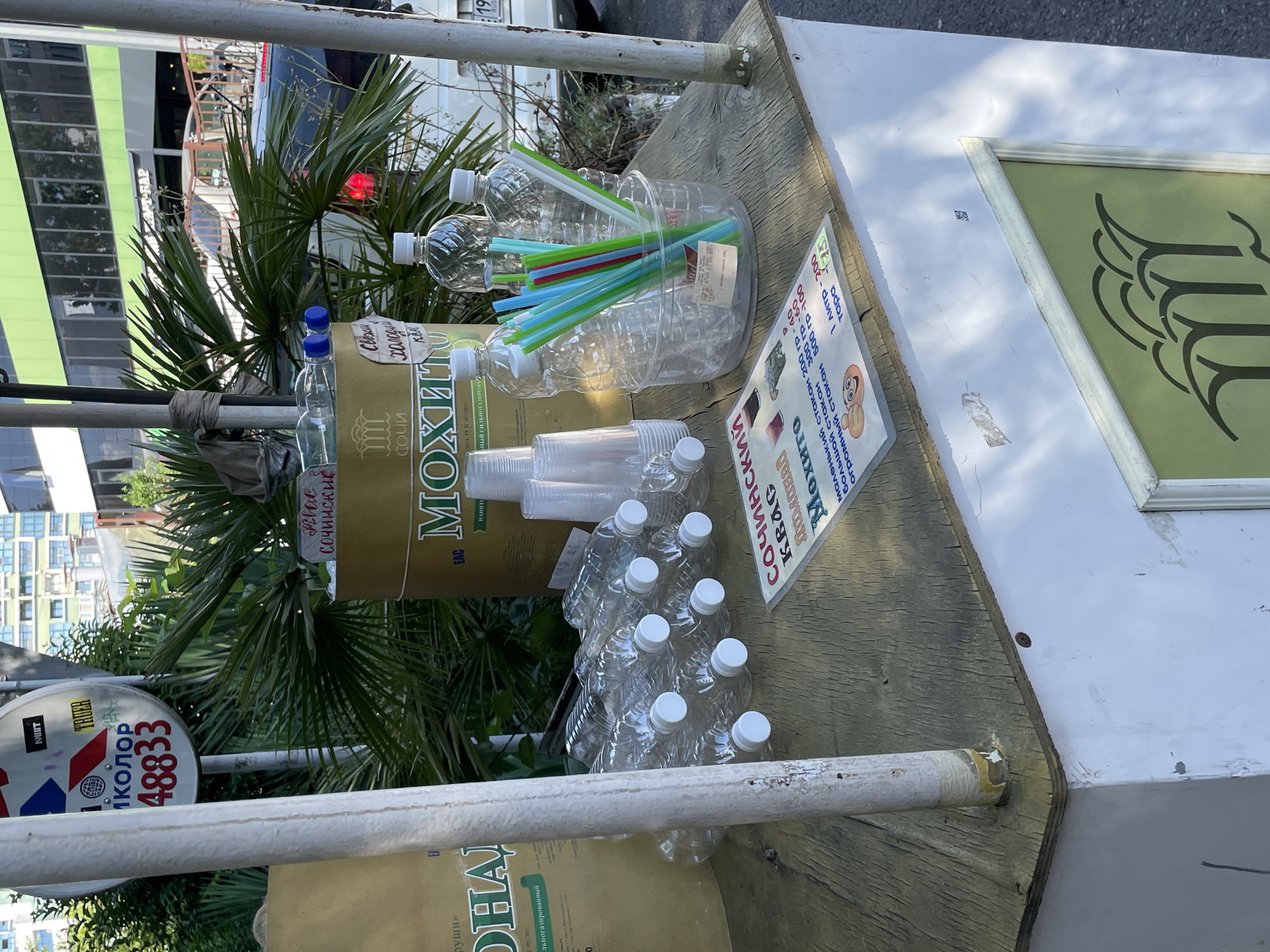 An outdoor beverage stand is set up on a sidewalk in Sochi, Russia, likely near a building with a green facade and palm trees. The stand is constructed from a white base topped with a weathered wooden surface. On the surface, several empty plastic water bottles are arranged in rows, with their white caps facing upwards. A large metal can, labeled "МОХИТО" (Mojito) in Russian and featuring an emblem of the city of Sochi, is visible. In front of the can, a clear plastic container holds an assortment of colorful plastic straws. A folded menu or price list lies on the wooden surface, displaying prices for different sizes of mojito drinks and other beverages in Russian currency (rubles). The menu features illustrations of drinks and a thumbs-up emoji. A white pole, possibly part of the stand's structure or a nearby signpost, runs vertically through the scene. In the background, cars and people are visible, suggesting a public area. The overall scene depicts a typical street vendor setup in a warm climate.