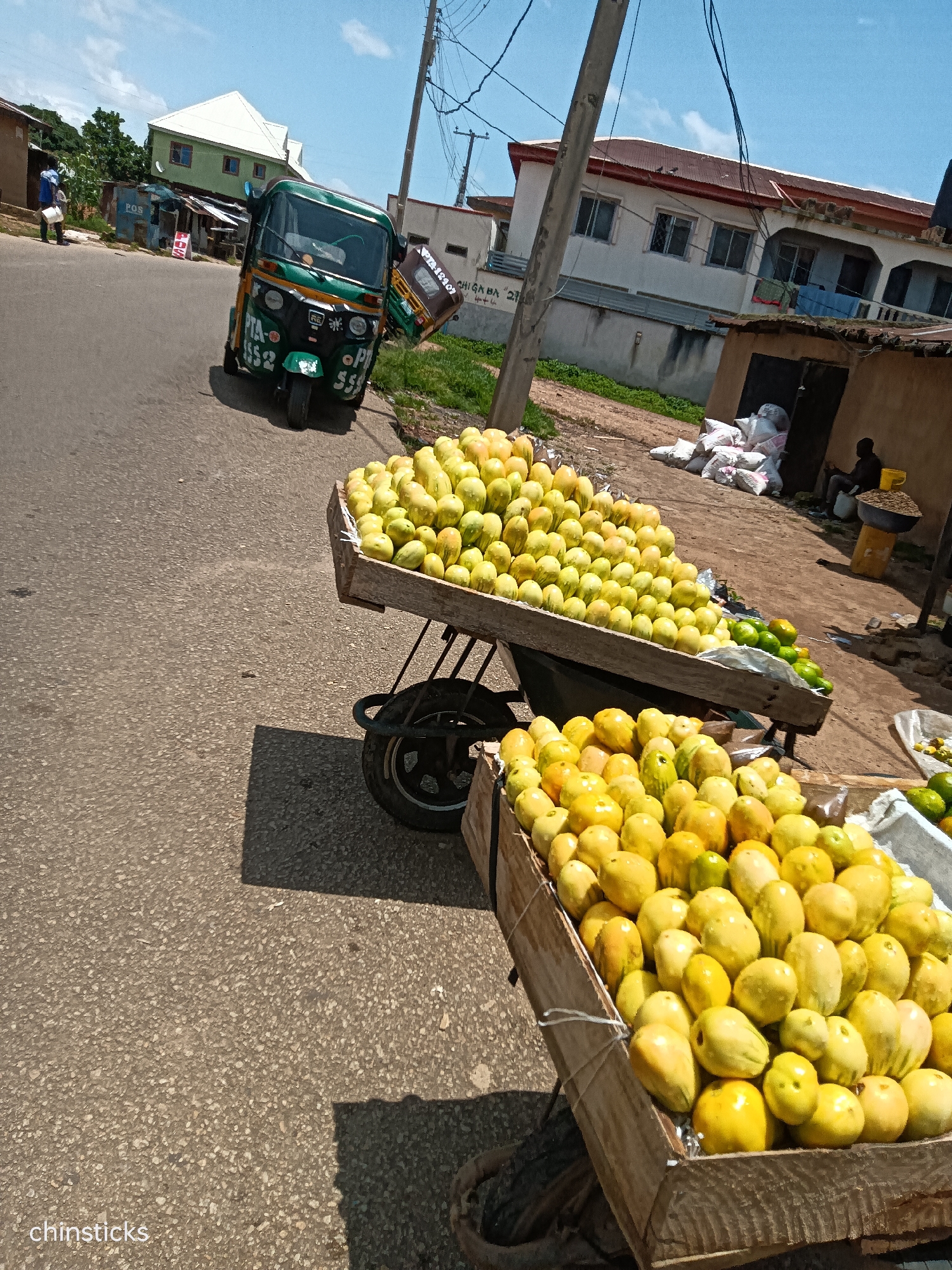 In the city of Jos, Nigeria, a street scene unfolds under a bright, sunny sky with scattered clouds. A green and yellow auto-rickshaw, identified by the license plate PTA-452-1552, is parked on the side of a paved road. In the foreground, two wooden carts are laden with a vibrant display of yellow fruits, likely mangoes, arranged in neat piles. Behind the fruit carts, a person is seated next to a collection of white sacks and a small stall. Buildings with red roofs and cream-colored walls line the street, some appearing residential and others commercial. A utility pole with numerous wires stands prominently on the right side of the image. The overall impression is that of a bustling local market on a typical day.