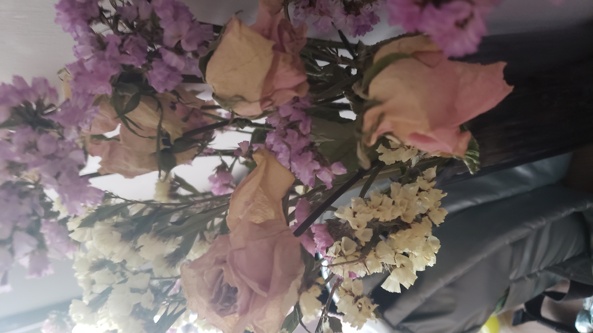The image shows a close-up of a bouquet of dried flowers, primarily roses and statice, in shades of pale pink, yellow, and lavender. The flowers appear to be in a vase, with some stems visible. The background is blurred, showing what appears to be a dark, possibly leather surface to the left, and a lighter wall to the right. The lighting suggests an indoor setting. There is no identifiable information within the image that would place it in Bratislava, Slovakia.