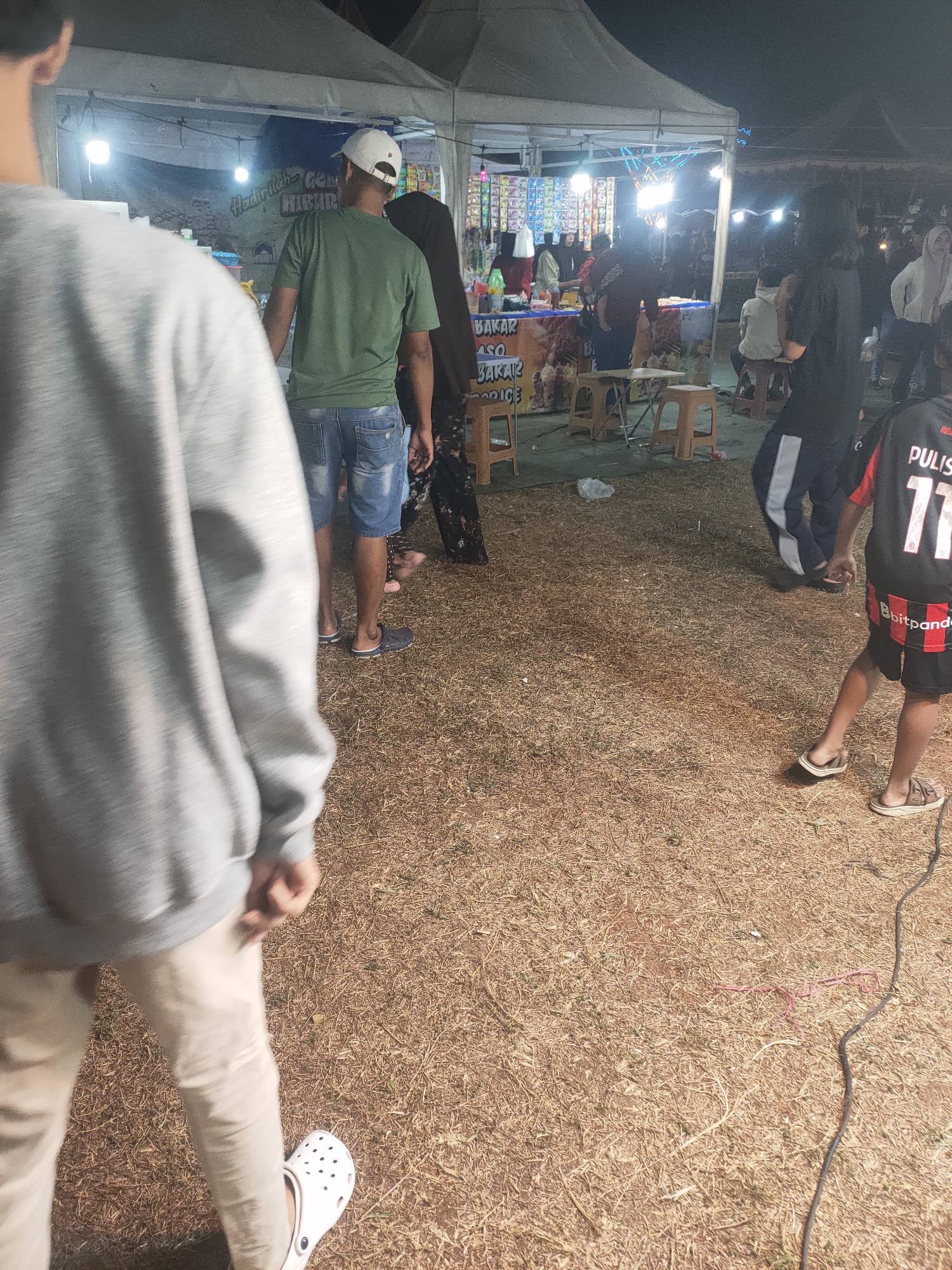 The image shows a nighttime scene at a street food stall or small market in Subang, Indonesia. In the foreground, the back of a person wearing a grey hoodie and light-colored pants is visible, with their left foot in a white Croc shoe. To the right, a child in a black and red soccer jersey with the number "11" and matching shorts is walking away from the camera, wearing sandals.
Behind them, a man wearing a green t-shirt and denim shorts stands near a stall, looking towards the interior. A white baseball cap is on his head. The stall itself has a banner with text that is partially obscured but appears to advertise food items like "BAKAR" (grill) and "ASO" (possibly referring to a type of dish). Various products are displayed on shelves behind the stall, illuminated by bright lights. Other people are visible in the background, some standing and some sitting at small tables. The ground is covered in dry grass and straw. Tents are set up overhead, suggesting an outdoor event or market. A dark cable runs across the ground in the lower right portion of the image.