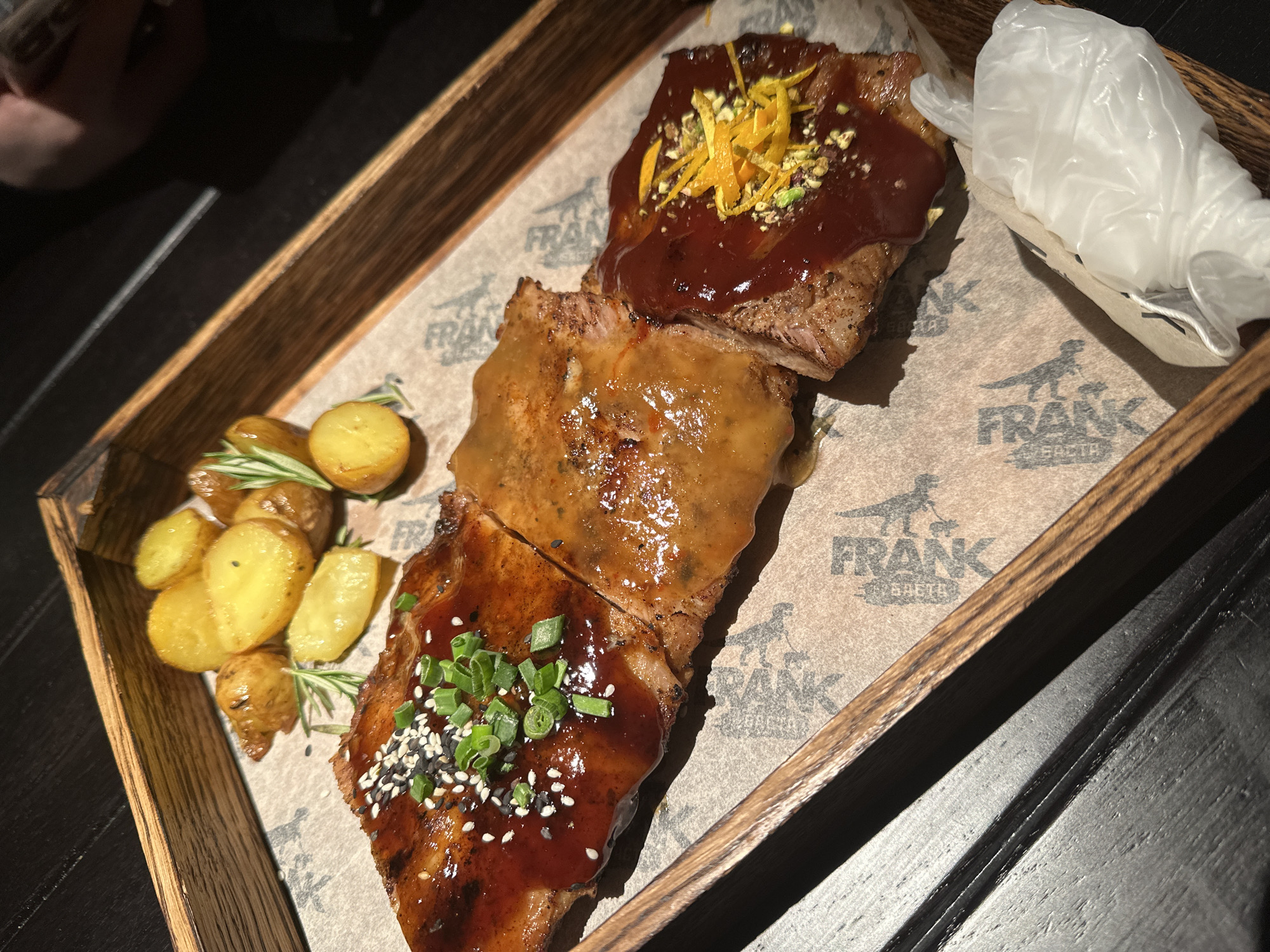 The image shows a wooden tray with three pieces of pork ribs covered in different sauces and toppings: one with a brown glaze topped with green onions and sesame seeds, one with a mustard-like sauce, and one with a dark red sauce topped with chopped orange zest and pistachios. A small pile of roasted potatoes with rosemary sprigs is on the left side of the tray. A pair of disposable plastic gloves is on the right. The tray is lined with paper printed with the name "FRANK," likely indicating the restaurant or establishment. While the exact location in Saratov, Russia, cannot be determined from the image, the tray's branding points to a possible association with a restaurant chain named "FRANK" known to have locations there.