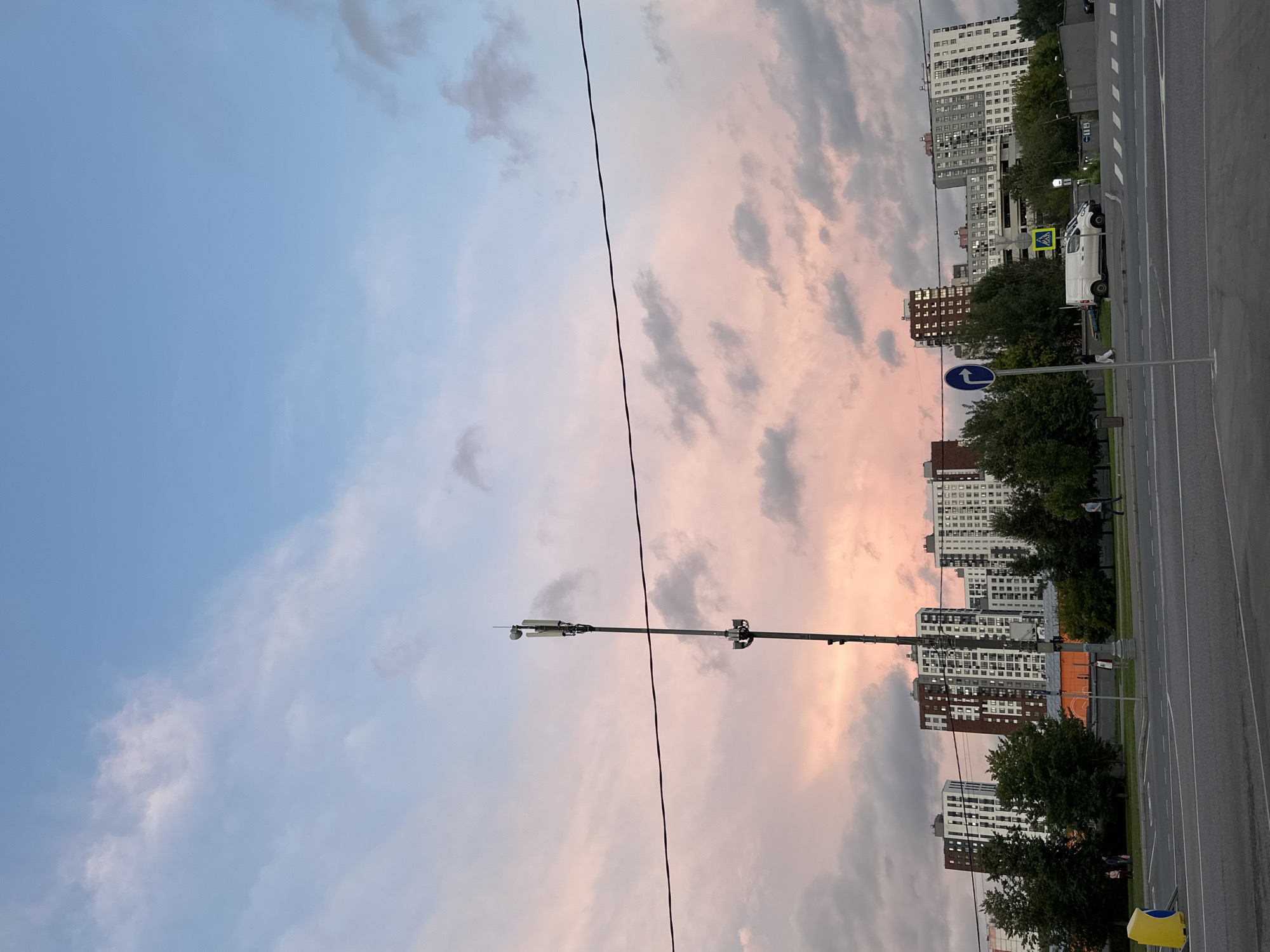 The image depicts a street scene in Moscow, Russia, during a twilight hour. The sky is a gradient of soft pink and blue, with scattered clouds. In the foreground, a multi-lane road with white lane markings is visible. A traffic light pole stands on the right side of the road.
In the middle ground, there is a sidewalk with several trees and a pedestrian crossing. A white van is parked on the sidewalk. A few individuals are walking along the sidewalk.
In the background, a row of tall, modern apartment buildings lines the horizon. A cell tower is prominent in the middle of the frame, extending towards the sky.
Overall, the scene conveys a sense of urban twilight, with the soft colors of the sky contrasting with the geometric forms of the buildings and infrastructure.