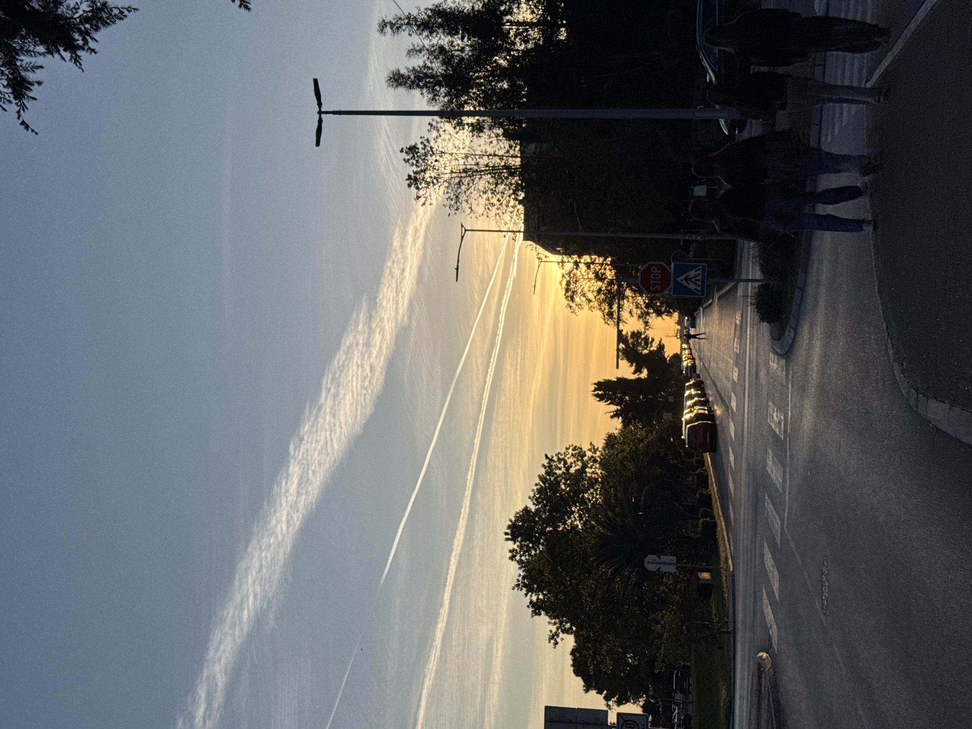 The image depicts a street scene in Bar, Montenegro, captured during either sunrise or sunset, indicated by the warm, golden light emanating from the horizon on the right. The sky transitions from a soft blue at the top left to a vibrant orange and yellow near the sun, adorned with two distinct, long airplane contrails stretching across the frame. One contrail is broad and feathery, while the other is thinner and more linear.
In the foreground, a paved road curves gently to the right, featuring a prominent zebra crossing. Several individuals are present, mostly silhouetted against the brighter background, suggesting they are either on or approaching the pedestrian crossing. One person appears to be pushing a stroller or cart. On the right side of the road, multiple cars are visible, including one red vehicle with reflections on its roof, indicating it's waiting or stopped at an intersection.
Lining the street are numerous trees with dense foliage, primarily seen in silhouette due to the backlighting, though some scattered sunlight illuminates their upper branches. Several streetlights, characterized by their two-lamp design, are positioned along the road and sidewalk. Visible road signs include a red octagonal "STOP" sign, a blue triangular pedestrian crossing sign, and a circular speed limit sign displaying "60" at the bottom right. The overall impression is a bustling urban environment bathed in the dramatic light of dawn or dusk.