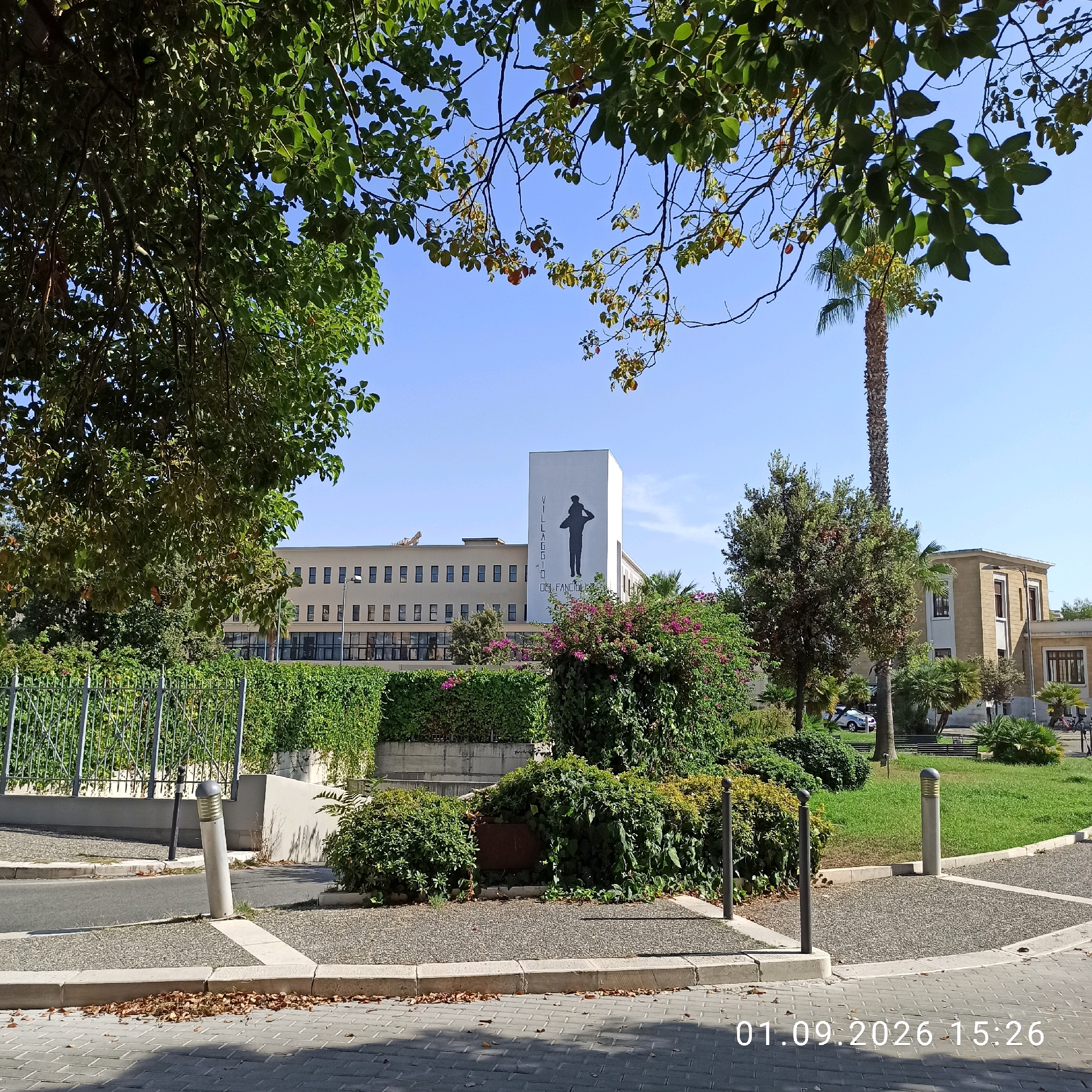 The image shows a bright, sunny day in Bari, Italy, with a view of the Villaggio del Fanciullo. In the foreground, a paved area with gray brickwork is visible, strewn with dry leaves. A curved sidewalk separates the paved area from a strip of gravel and then a grassy area. Several objects are present in the middle ground, including a tall, slender street lamp, a concrete planter, and various bushes and trees.
A prominent white rectangular building dominates the background, featuring a large black silhouette of a person on its facade. The building has multiple windows arranged in horizontal rows. To the left of this building is a longer, lower structure, also with numerous windows. Further to the right, a smaller, tan building with dark-trimmed windows is visible. A tall palm tree stands between the white building and the tan building. The sky is clear and blue. The timestamp "01.09.2026 15:26" is overlaid in the lower right corner of the image.