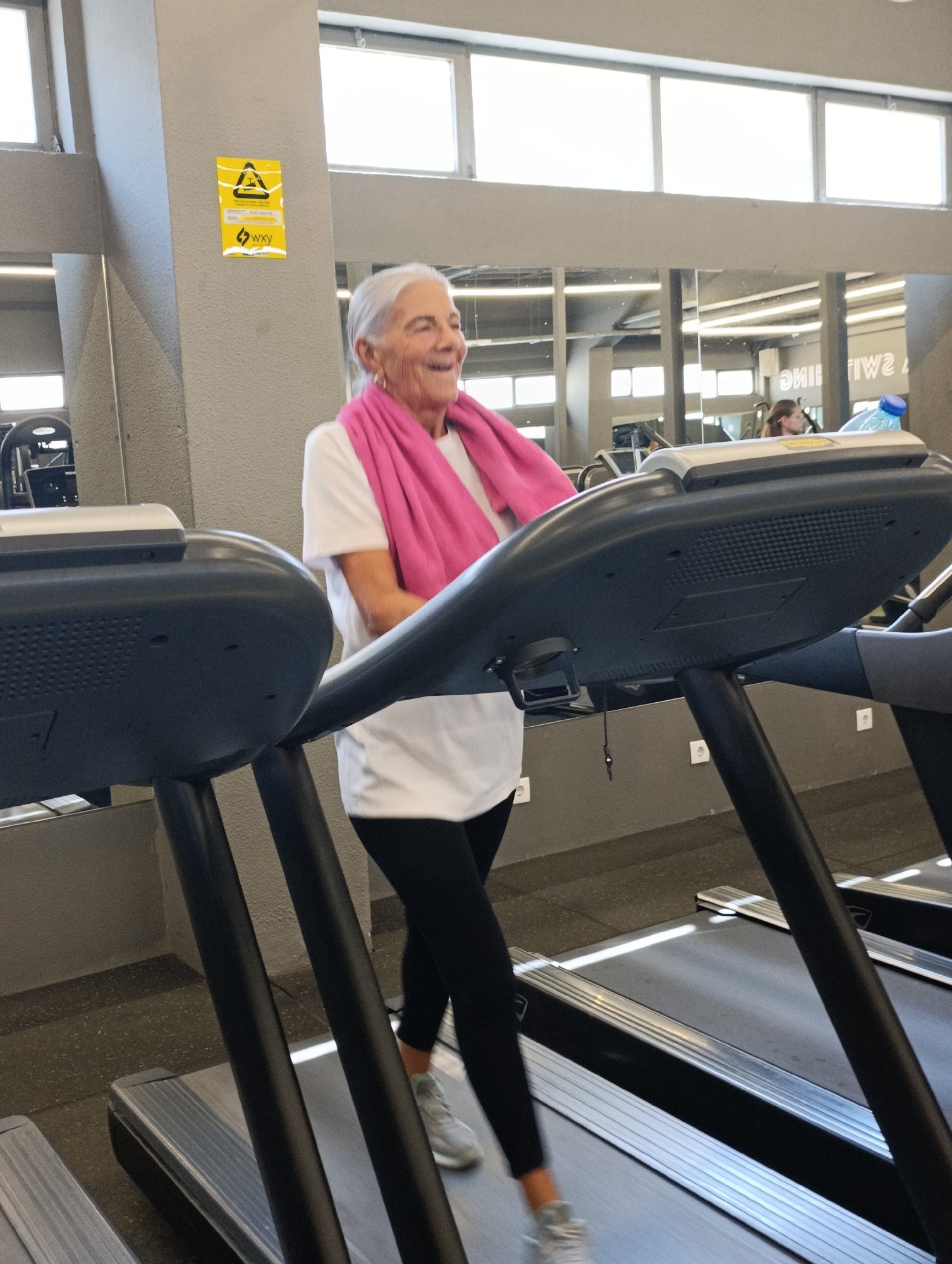 An elderly woman with gray hair and a pink towel around her neck is walking on a treadmill in a gym. She is wearing a white t-shirt and black leggings. The gym appears to have mirrors on the walls and other gym equipment in the background. There are windows on the ceiling and a sign on the wall with a yellow triangle. The overall scene suggests a fitness facility. The location is unknown but the presence of a "WXY" sticker and a specific type of window may indicate it's in Setúbal, Portugal.