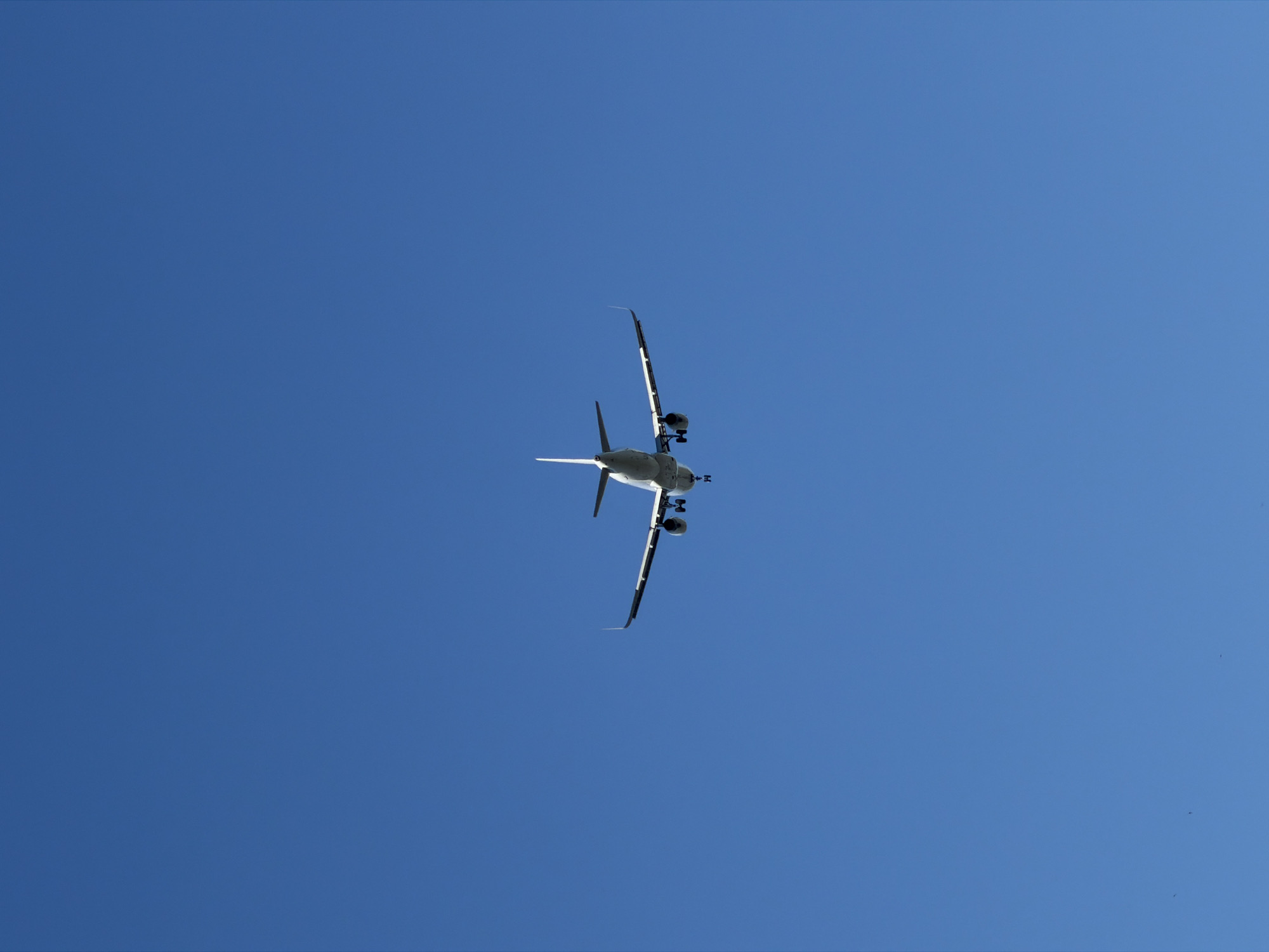 An airplane is seen from below, flying against a clear blue sky. The aircraft is white with wings extended and its landing gear deployed, suggesting it is either landing or taking off. The image does not contain any information that would allow for the determination of its location within Burgas, Bulgaria.