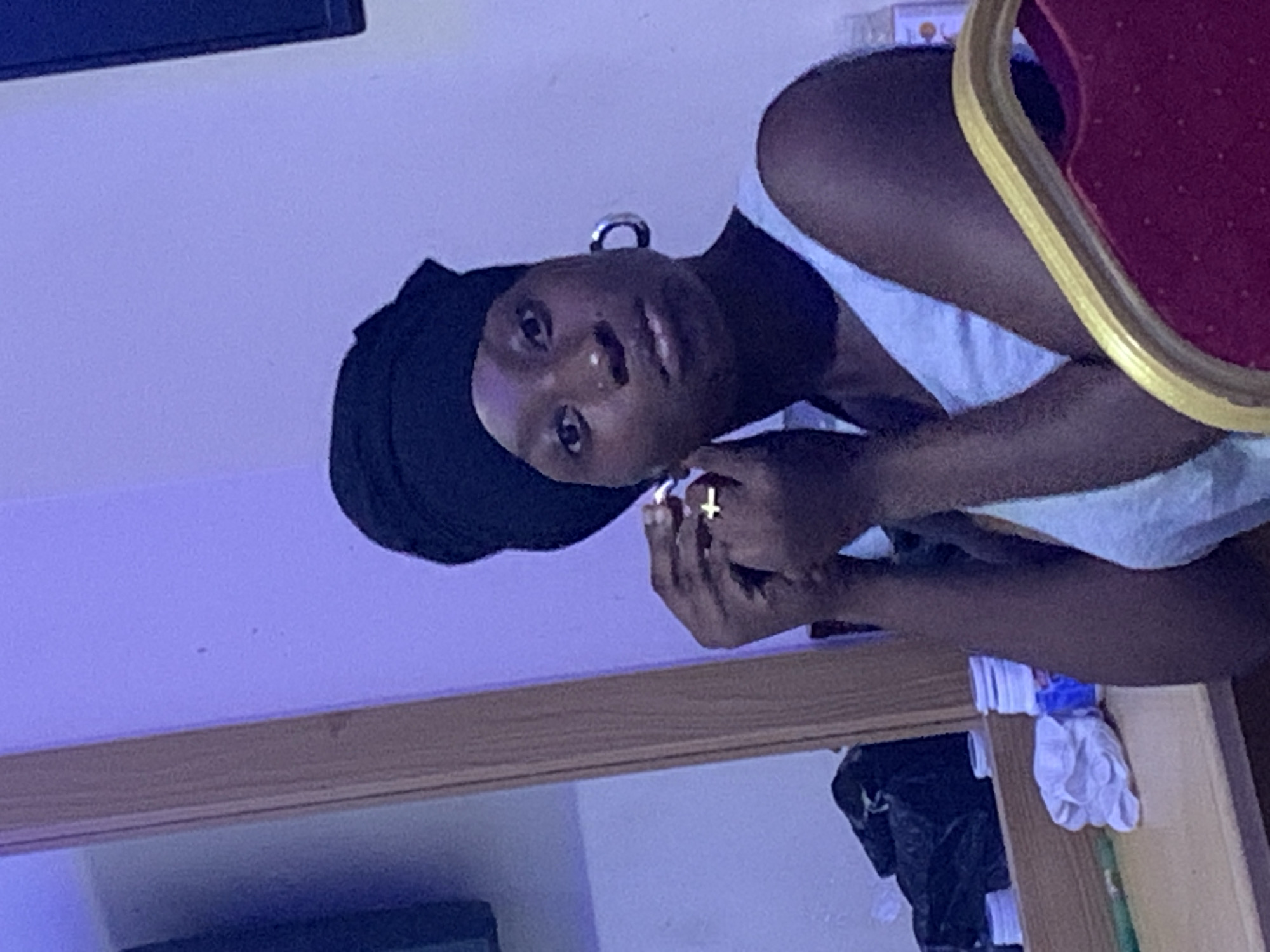A dark-skinned person with a black headscarf and large hoop earrings is the primary subject of the image. They are wearing a light-colored top. A gold cross ring is visible on one of their fingers.
The background appears to be an interior space with a mirror reflecting items, including a black bag and some white objects.
Location: Not possible to determine the specific location in Lokoja, Nigeria.
