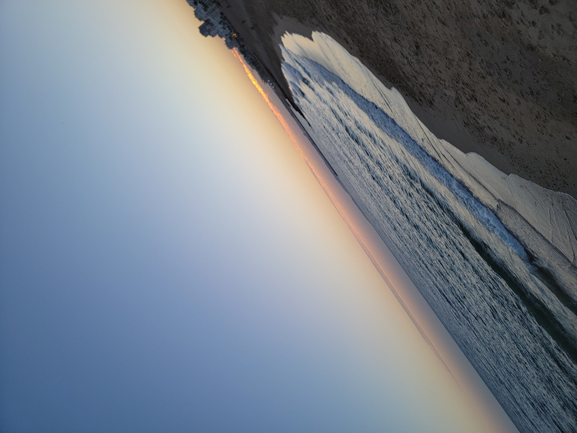 A wide-angle, low-angle, shot of a diagonal beach scene during dusk in Quarteira, Portugal. The image is dominated by the gradient sky, transitioning from a clear, light blue at the top left to a soft orange and pink hue near the horizon. The ocean is on the left, appearing dark blue with white crests of waves rolling onto the sandy beach. The beach occupies the right side of the frame, appearing dark brown with visible footprints in the sand. In the background, a line of buildings, likely apartments or hotels, can be seen along the coast, illuminated by the fading sunlight. A few small, dark figures, presumably people, are visible on the beach and near the buildings, but their details are indistinct. The overall impression is of a peaceful evening by the sea.