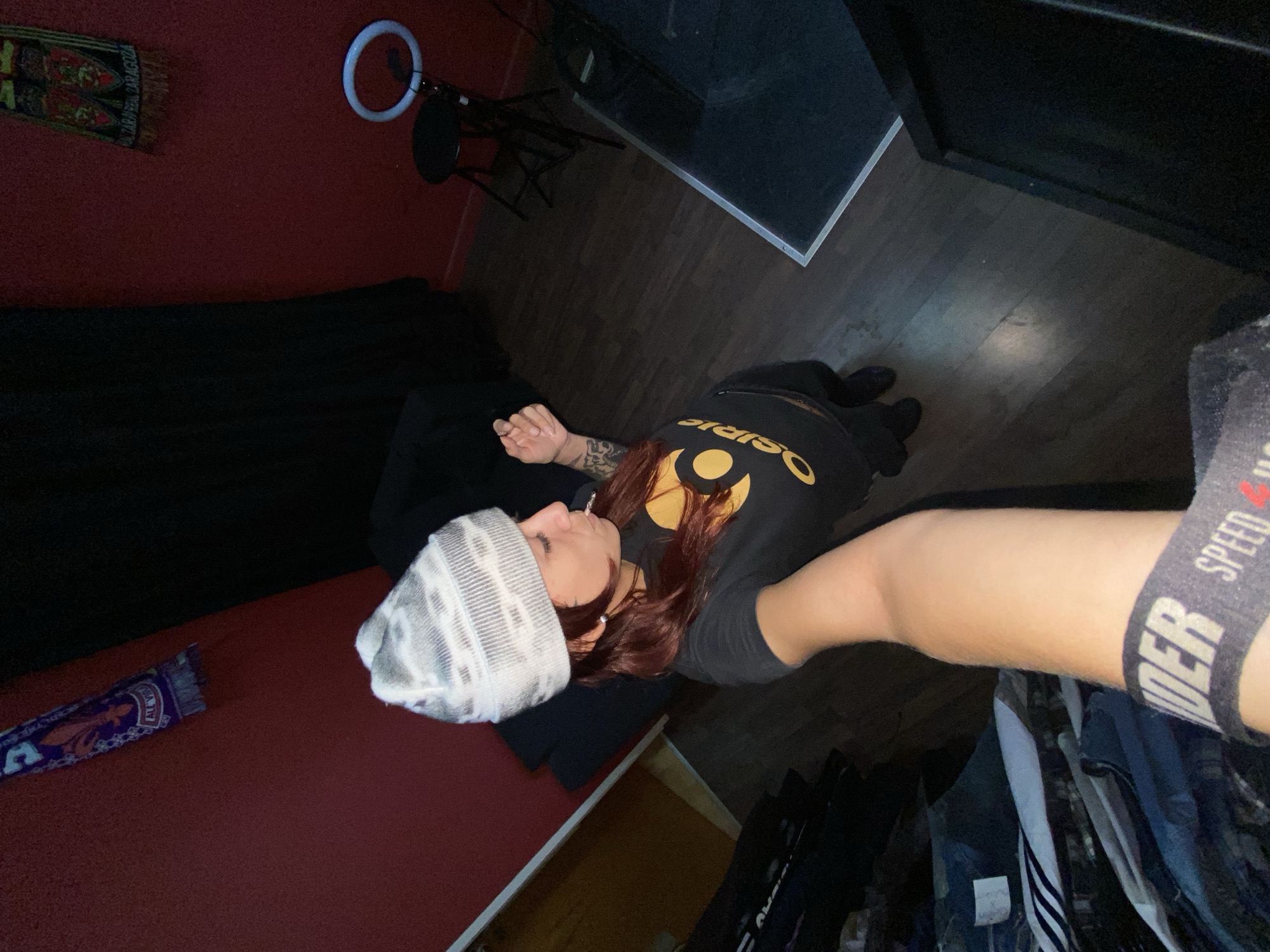 A low-angle, indoor shot depicting a person in a black t-shirt with gold lettering lying down. The person wears a gray and white beanie. Red hair is visible. The arm closest to the camera reveals an "UNDER" underwear band. The surrounding environment includes a red wall, a black blanket, wooden floor, a clothing rack with hanging clothes, a ring light and microphone stand. The location is likely a bedroom or studio in Llavallol, Argentina.