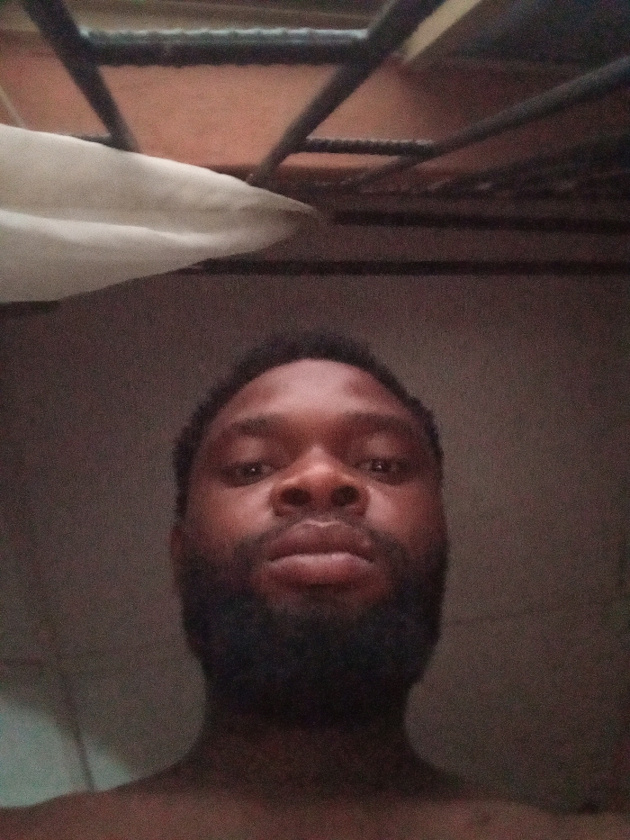 An upward-angle close-up photograph depicts an adult male, likely of African descent, centered in the frame. He has dark skin, short dark hair, and a full, dark beard. His eyes are open and directed towards the camera, with a neutral expression. Only his head, neck, and upper chest are visible; no specific clothing is discernible. The background is dimly lit and out of focus. Above his head, a structure composed of dark, intersecting metal bars is visible against a lighter, possibly orange-brown, ceiling or surface. To the upper left, a portion of a white, folded or draped fabric object is present. The overall lighting is low, contributing to a grainy image quality. The scene appears to be an indoor setting in Magboro, Nigeria.