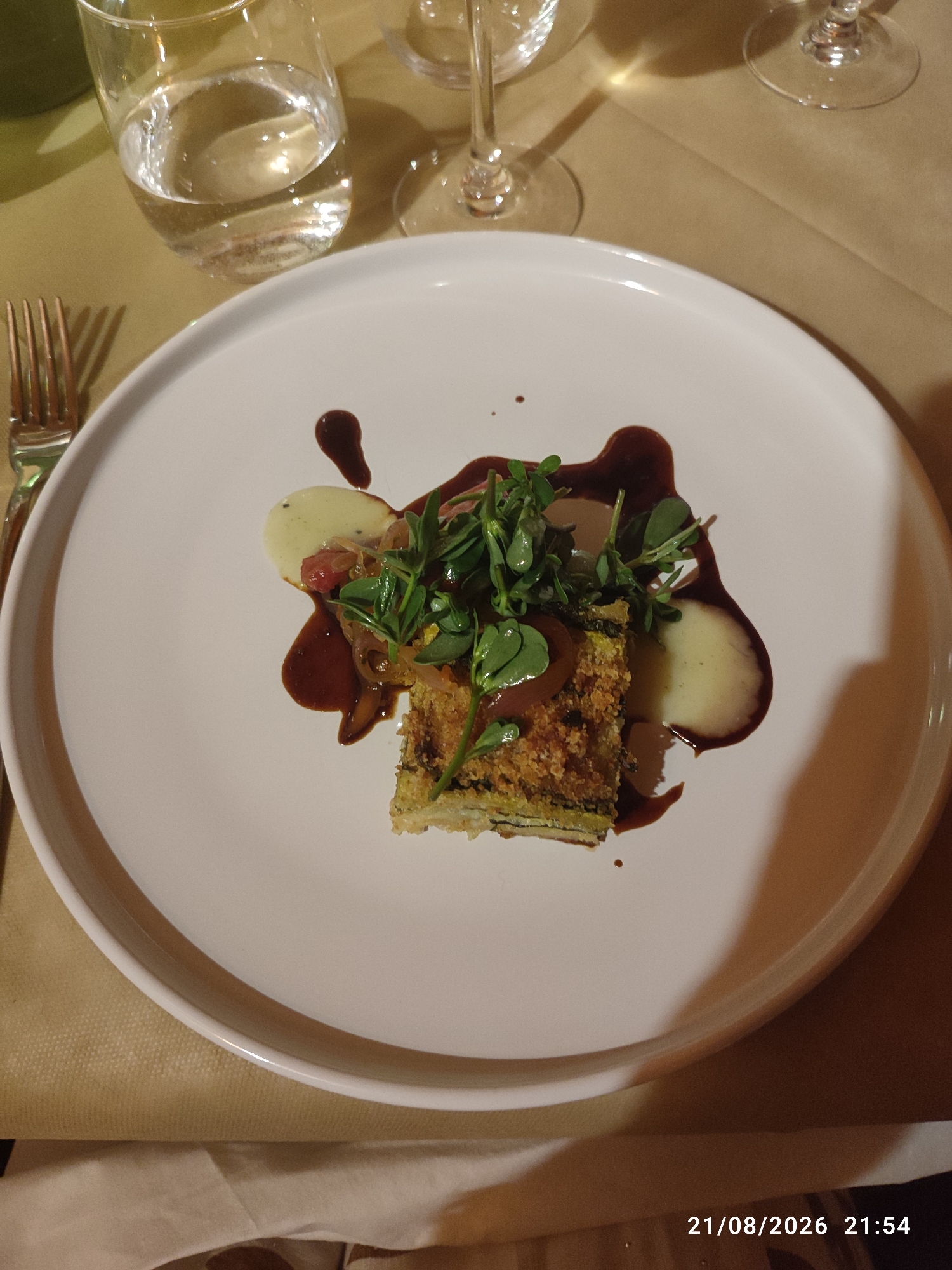 A close-up, eye-level shot shows a white, circular plate containing a piece of layered food, possibly a gratin or terrine, with a breadcrumb coating. The food is garnished with fresh green herbs and topped with thinly sliced red onions. A creamy, pale sauce is dolloped on the plate, and a dark, rich sauce is artistically drizzled around the food. To the left of the plate, a fork with four tines is visible, and beyond that, a partially filled glass of clear liquid. In the background, another glass stem is visible. The table is covered with a light brown, textured cloth, and a white napkin is partially visible in the foreground. The lighting is dim and warm, creating shadows. The scene is likely a restaurant setting. The timestamp "21/08/2026 21:54" suggests this image was taken on August 21, 2026, at 9:54 PM. The image does not provide any specific information about the location in the city of Militello in Val di Catania, Italy.