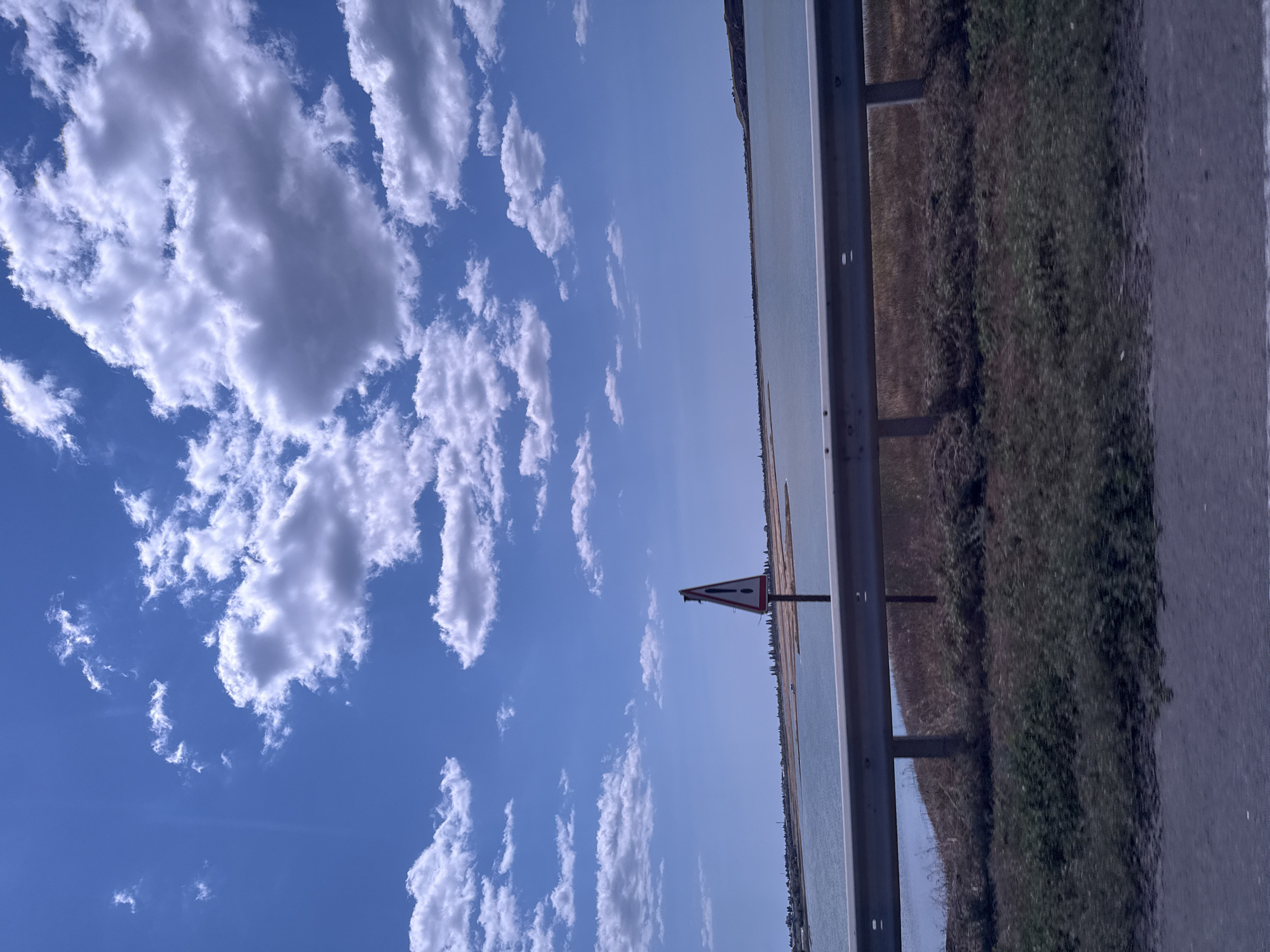 This image captures a roadside view of a body of water, possibly a lake or estuary, under a bright blue sky filled with scattered white clouds. In the foreground, a metallic guardrail runs horizontally across the frame, separating the road from the grassy embankment and the water beyond. A warning sign, triangular with a red border and a black exclamation mark on a white background, is prominently placed on a pole to the left of the guardrail. The water is a light blue-green color, and the distant shore is lined with trees and appears to have some structures. The overall impression is of a scenic, perhaps slightly remote, location on a clear day. The image was taken from a moving vehicle, as indicated by the slight motion blur in the foreground. There is no indication that this location is in Koshary, Ukraine.