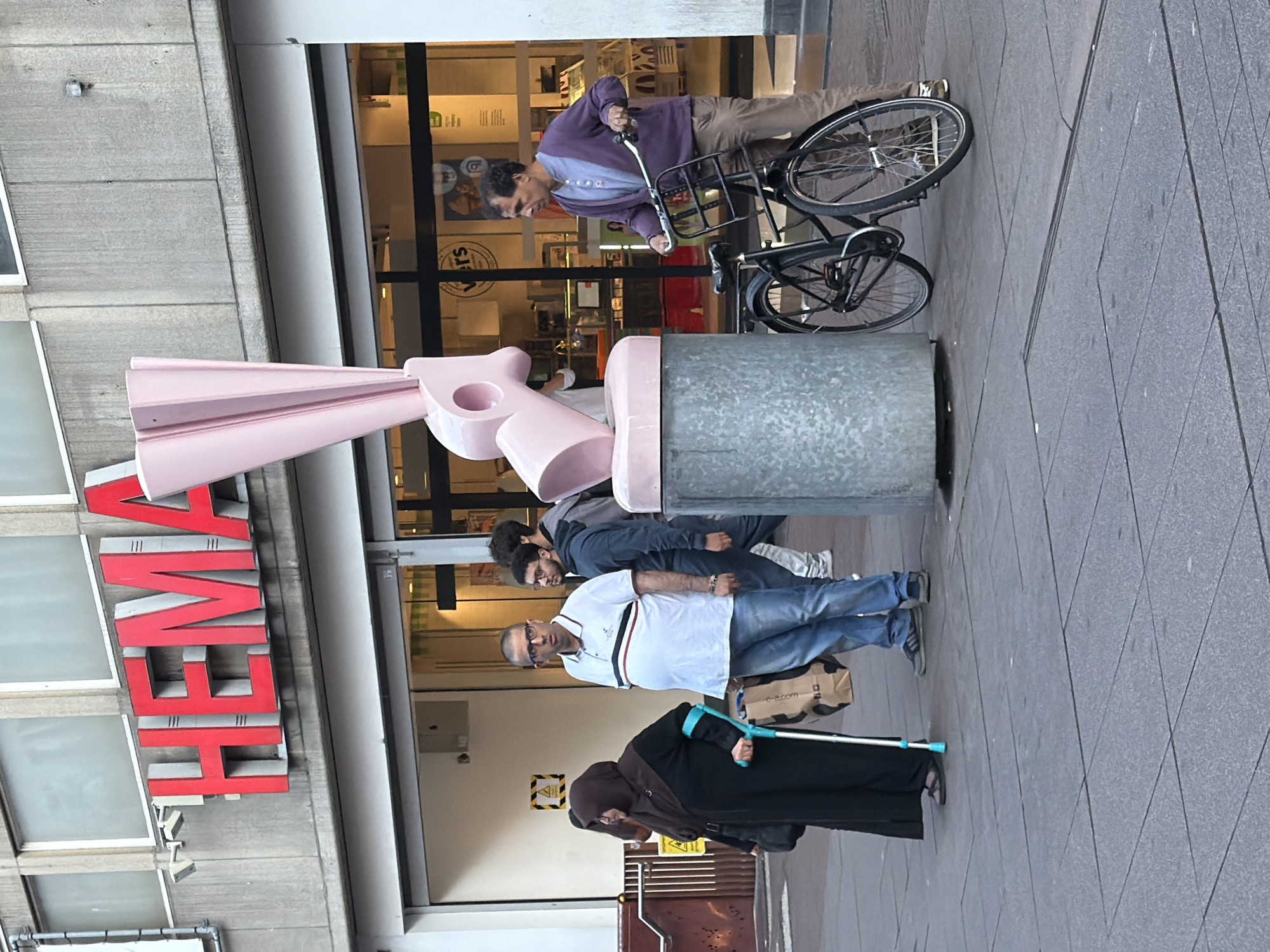A man in a purple cardigan and brown pants is standing next to a black bicycle. He is looking to his right. Behind him, a woman wearing a black abaya and headscarf is walking with a crutch. She is looking down. To her right, a man in a white polo shirt and blue jeans is standing and looking to his left. Behind them, the red HEMA logo is visible on a building. The scene appears to be a city street in The Hague, Netherlands.