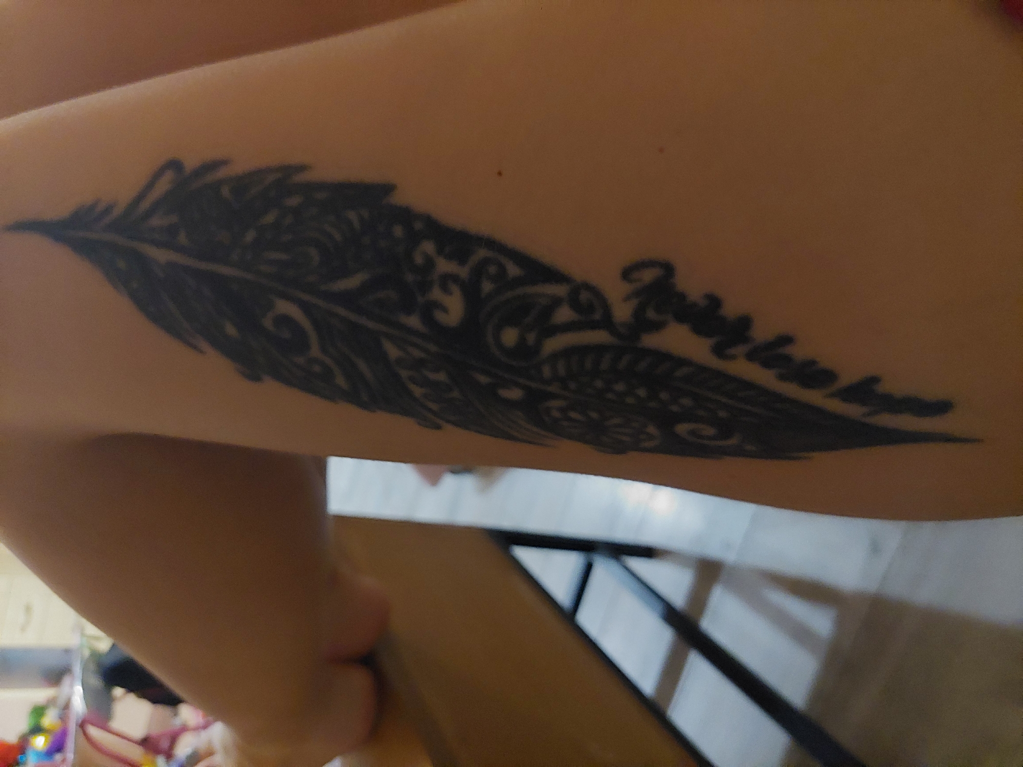A close-up shot shows a tattoo of a feather on a person's thigh. The feather tattoo is black and intricate, with detailed patterns. Below the feather, some faint text is visible, appearing to be written in cursive. The person's skin tone is fair. The background is blurred, but suggests an indoor environment with a wooden surface in the foreground and a light-colored floor with shadows. No information about the city of Plovdiv, Bulgaria is present in the image.
