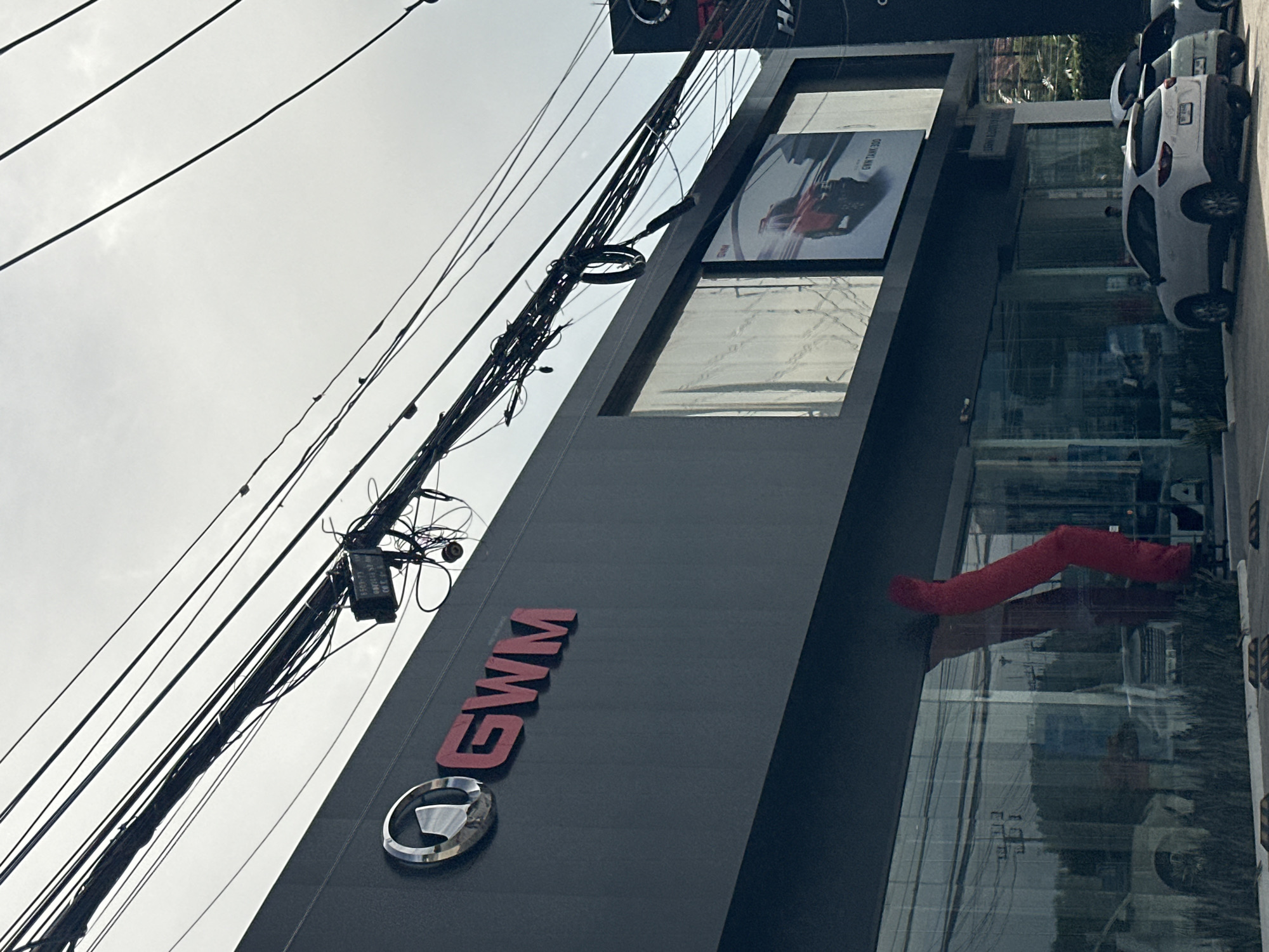The exterior of a GWM car dealership is visible, with the GWM logo and red lettering prominently displayed on the dark grey facade. A large digital billboard on the building shows an advertisement for a red GWM TANK 300. The building features large glass windows, reflecting the surroundings, and grass is planted along the base. A bright red inflatable tube dancer is positioned in front of the glass, its form bent and distorted. Several cars are parked outside the dealership, including a white SUV. Above the building, a dense network of electrical wires crisscrosses against a bright, overcast sky. The overall scene suggests a commercial establishment in an urban or suburban setting, likely in Kathu, Thailand, given the context.
