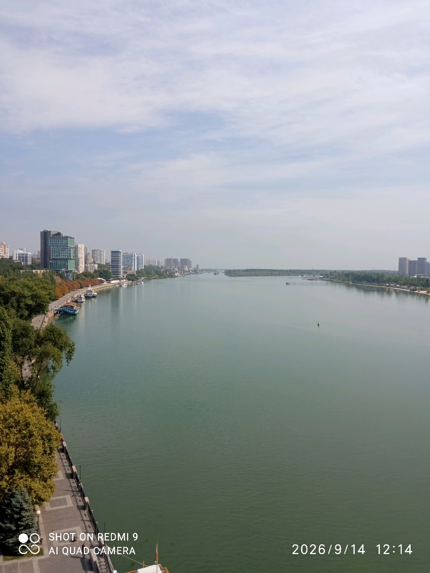 This is an eye-level, long shot photograph of the Don River in Rostov-on-Don, Russia, on September 14, 2026, at 12:14 PM. The river dominates the scene, its murky green water stretching towards the horizon under a hazy, overcast sky. On the left bank, a promenade lined with trees and walkways runs along the water's edge. Several buildings of varying heights, including modern high-rises and older structures, are visible behind the trees, forming the city skyline. A few boats are docked along the promenade. On the far bank of the river, more buildings and industrial cranes are discernible, along with a treeline. A small green navigational buoy is visible in the middle of the river. The bottom left corner of the image contains a watermark indicating it was taken by "SHOT ON REDMI 9 AI QUAD CAMERA."