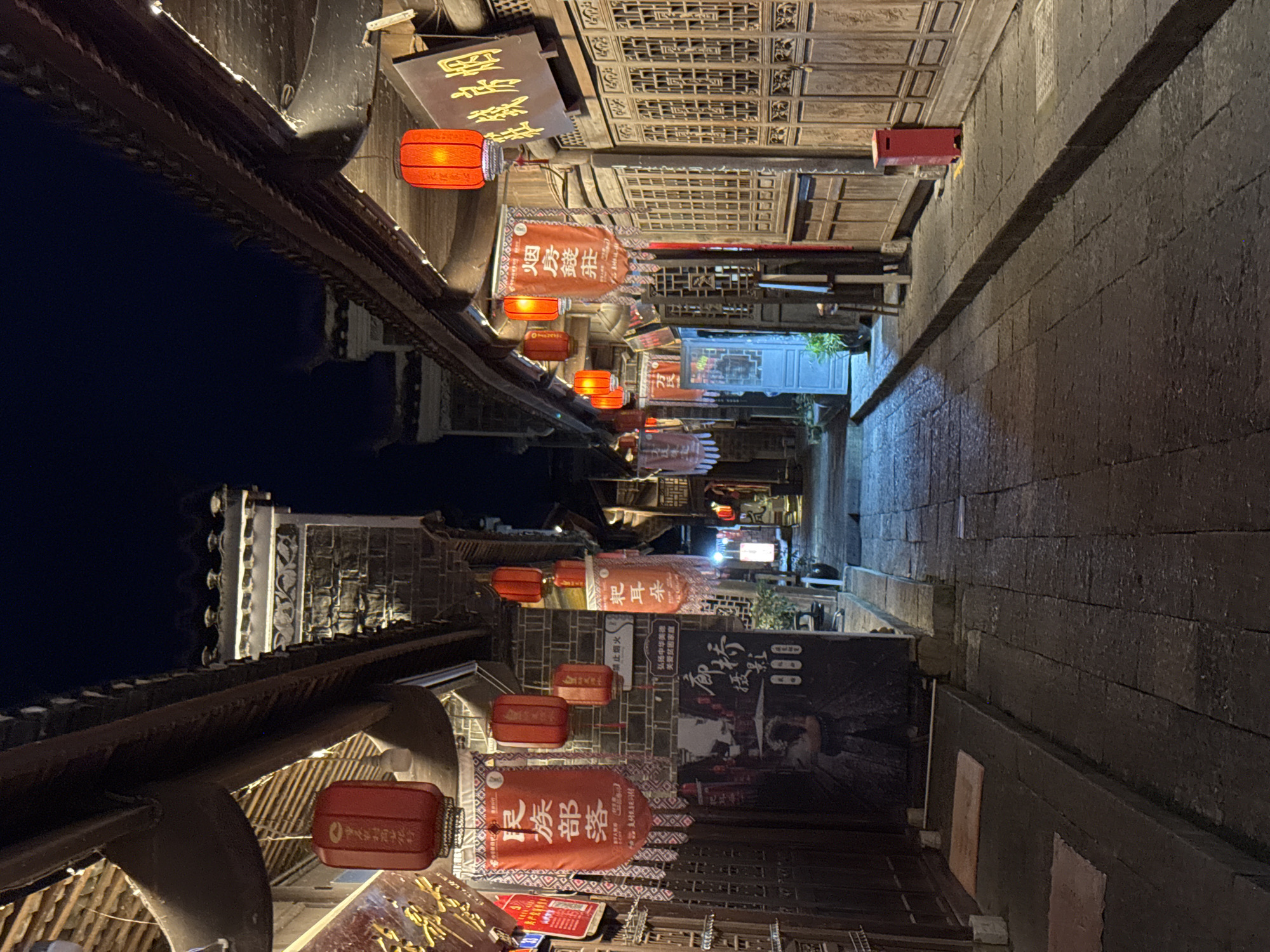 Here's a detailed description of the image:
**Scene Overview:** The photograph depicts an alleyway at night in what appears to be an old district, likely in Zhuoshui, China. The perspective is looking down the narrow street, with buildings flanking either side.
**Buildings:**
* The buildings are of traditional Chinese architectural style, featuring dark wood, intricate latticework, and overhanging roofs.
* The buildings on the left side appear to face a body of water, possibly a canal. Their reflection is faintly visible on the water's surface.
* Buildings along the right side have a stone-wall appearance.
**Lighting and Decorations:**
* Red lanterns are strung along the alleyway, providing the primary source of light.
* Banners with Chinese characters hang from the buildings, adding to the cultural ambiance. The banners contain Chinese text.
* The lighting adds an air of warmth and vibrancy to the scene.
**Objects and Signs:**
* There are signs with Chinese characters hanging on the buildings.
* Advertising posters and various small items are affixed to the walls.
**Environment:**
* The alleyway is paved with stone or brick.
* The overall environment is somewhat dimly lit, but the lanterns provide focal points of illumination.
**Overall Impression:** The scene is evocative of a historic Chinese town, likely a tourist destination in Zhuoshui, China. The combination of traditional architecture, lighting, and cultural decorations creates a visually rich and atmospheric setting.