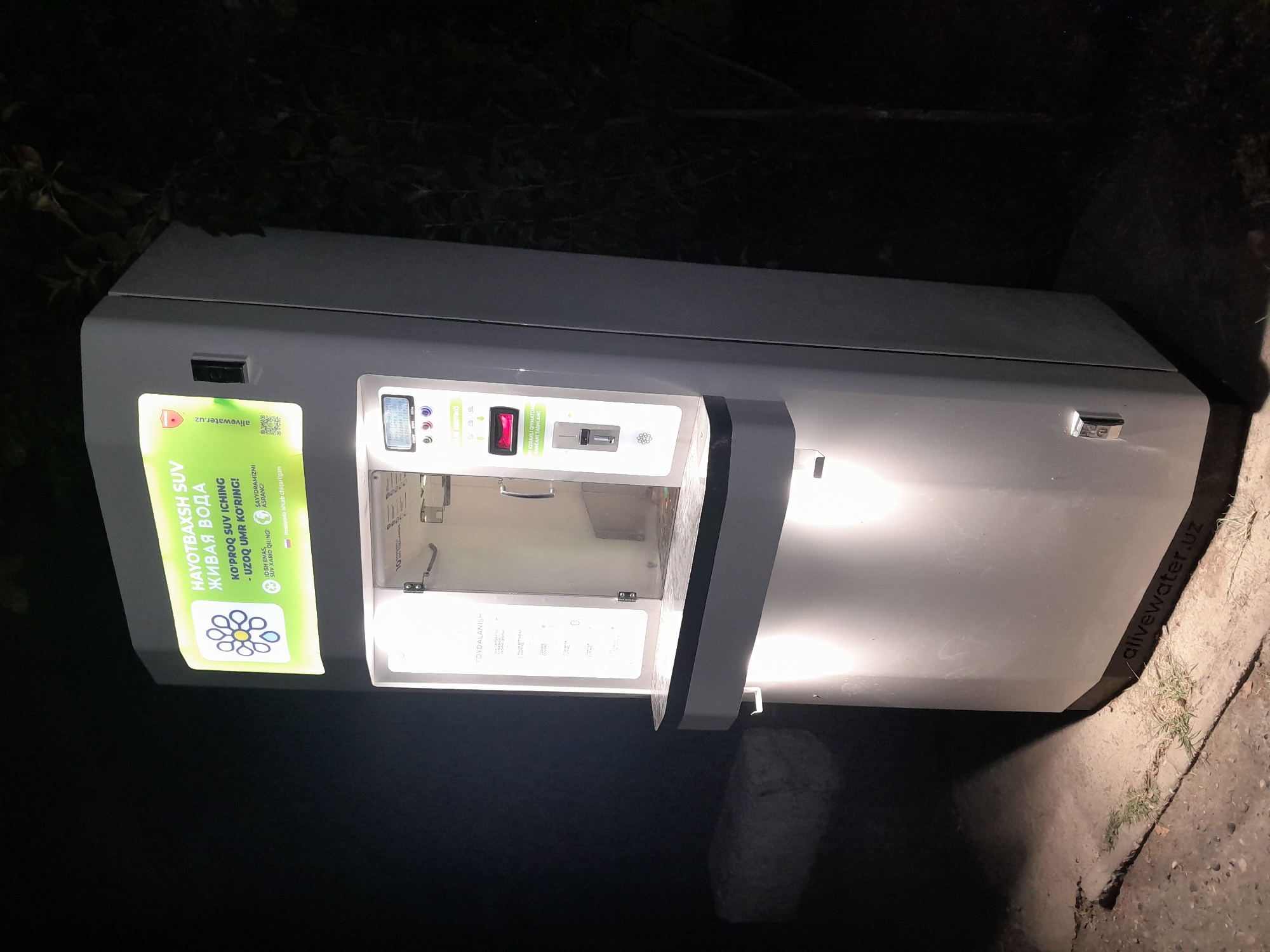 The image shows a water vending machine at night in Fergana City, Uzbekistan. The machine is lit from within, with a bright sign at the top that reads "HAYOTBAXSH SUV" and "ЖИВАЯ ВОДА" in Uzbek and Russian respectively. Below this, it says "KO'PROQ SUV ICHING - UZOQ UMR KO'RING!" which translates to "DRINK MORE WATER - LIVE LONGER!". The machine has a touch screen interface and a dispensing slot. A QR code and a website address "alivewater.uz" are also visible on the sign. The machine is situated on a paved area, with some grass growing through cracks in the pavement. A large, light-colored rock is visible in the background. Trees are present in the dark background, indicating an outdoor location. The overall lighting suggests it is nighttime and the machine is the primary light source.