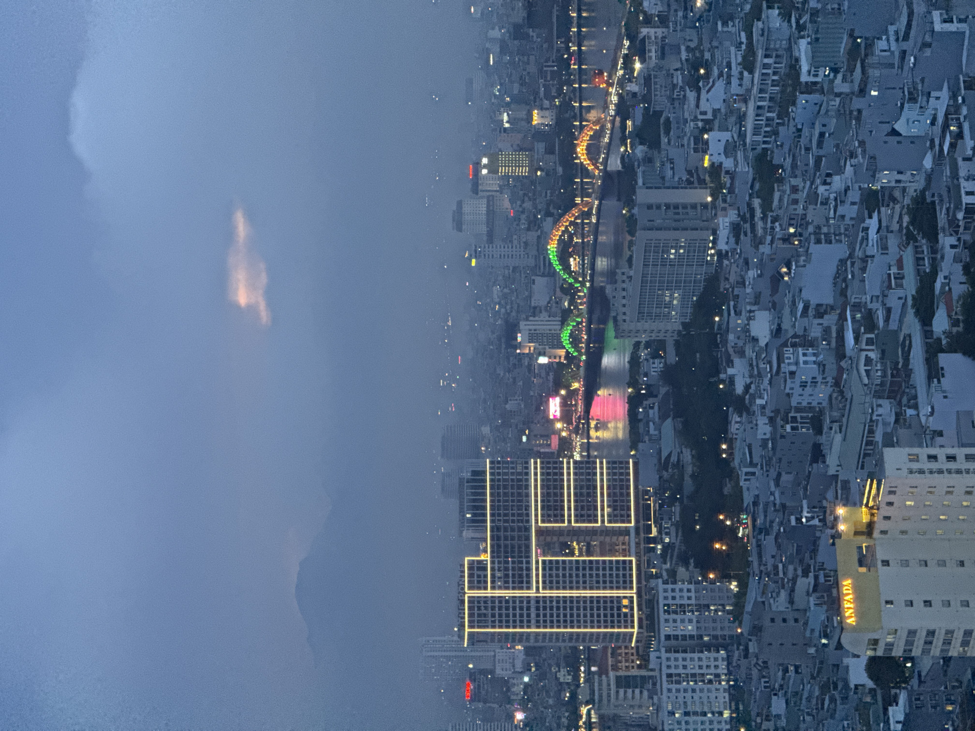 This is a high-angle, vertical, medium shot of a city skyline at dusk, likely in Da Nang, Vietnam. The sky is a hazy blue with some lighter clouds, one of which is tinged with pink. In the background, faint mountain silhouettes are visible through the atmospheric haze. The city is densely populated with numerous buildings of varying heights, many of which are illuminated with lights.
A prominent, modern building in the mid-left of the frame is illuminated with a distinctive pattern of yellow lights, giving it a geometric, almost "H" shape. The name "THE POINTE" is visible on its rooftop. To the right of this building, a bridge crosses a body of water, and it is lit with colorful lights, appearing green and yellow. The river or bay reflects the lights of the bridge and the city.
In the foreground, a building with the illuminated orange text "ANFADA" is visible. The overall scene depicts a bustling urban environment transitioning from day to night, with a clear view of the city's architecture and illuminated features. No people are discernable in the image. The atmosphere suggests a humid or hazy environment, common in coastal cities.