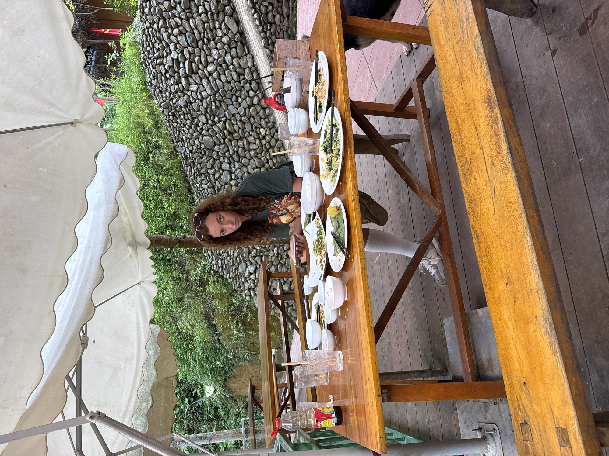 A young woman with curly brown hair and sunglasses perched on her head sits at a wooden table outdoors, looking at her phone. She is wearing a green t-shirt with a chicken design and white socks. The table is covered with dishes, bowls, glasses, and bottles of condiments. A dog can be seen partially behind the table on the right. The scene appears to be a restaurant or outdoor dining area in a rural setting, possibly Cổ Lũng Commune, Vietnam, with a stone wall and lush greenery in the background.