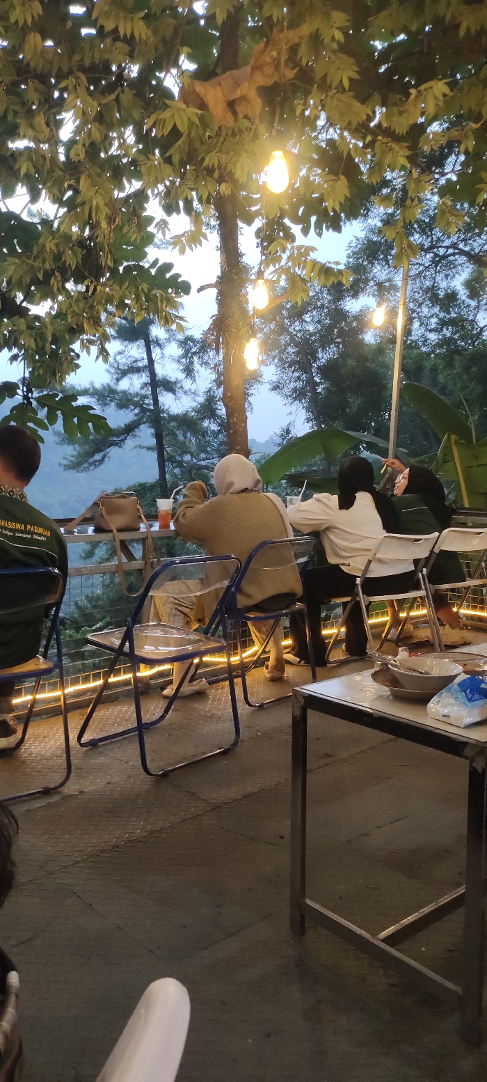The image depicts an outdoor, elevated cafe or restaurant setting in Cembor, Indonesia, during twilight or evening hours. Four individuals are visible, seated at tables positioned on a metal-plated floor with a railing overlooking a forested valley.

In the foreground, the back of a person wearing a dark green top with "MAHASISWA PASURUAN" text is visible on the left, seated in a clear plastic chair with a blue metal frame. To their right, a person in a light brown knitted top and light-colored headscarf (hijab) is seated facing away, leaning towards a clear plastic cup with a straw on a small metal table. A brown shoulder bag rests on this table. To the right of this person, two individuals are seated at a separate table. One wears a white top and a dark headscarf; the other wears a dark green top and a dark headscarf, appearing to engage in conversation, with one holding a thin object to their mouth. These two are seated on white folding chairs with metal frames.

In the immediate foreground on the right, a rectangular metal table holds two white bowls, one containing cutlery (fork or spoon), and a clear plastic bag. Overhead, string lights with glowing Edison-style bulbs are strung between large, leafy trees, providing artificial illumination. More string lights are visible along a railing or structure in the mid-ground. The background features dense, dark green foliage, including pine-like trees, extending down into a misty or hazy valley, indicating a natural, elevated landscape. The sky above the trees is a soft, pale blue-gray, consistent with dusk.